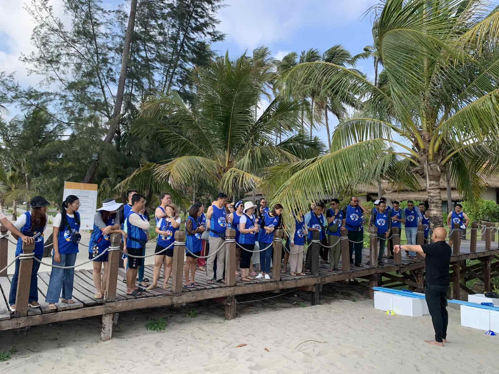 Tour group wearing matching blue shirts listening to guide on wooden boardwalk near palm trees.
