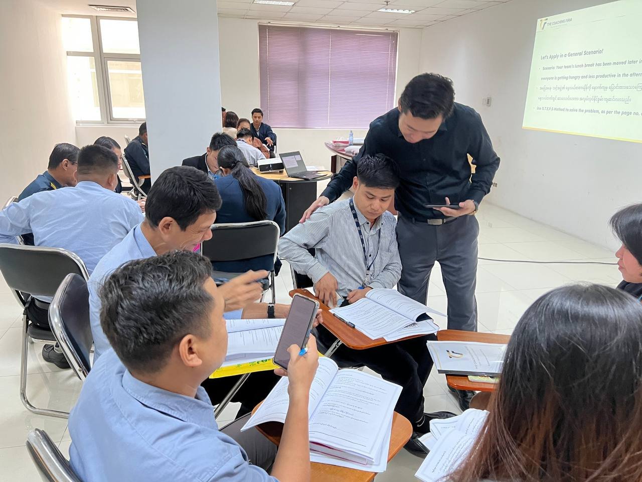 A group of professionals in a classroom or conference room engaged in a discussion, working with documents, laptops, and smartphones.