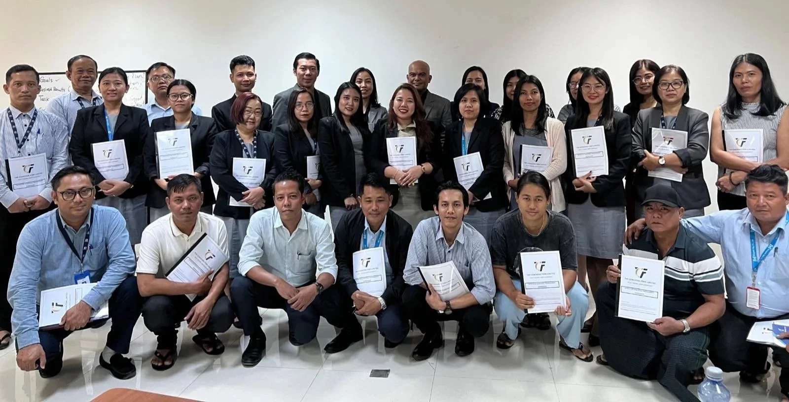 Group of men and women in business attire holding certificates, standing and kneeling in a conference room.