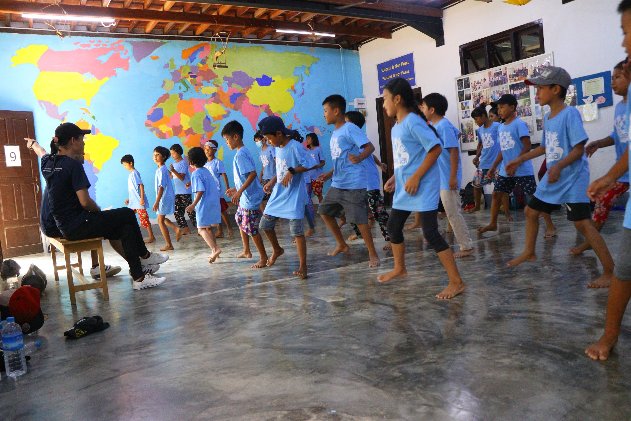 Group of children in matching blue t-shirts participating in an indoor activity, with a colorful world map mural on the wall behind them.