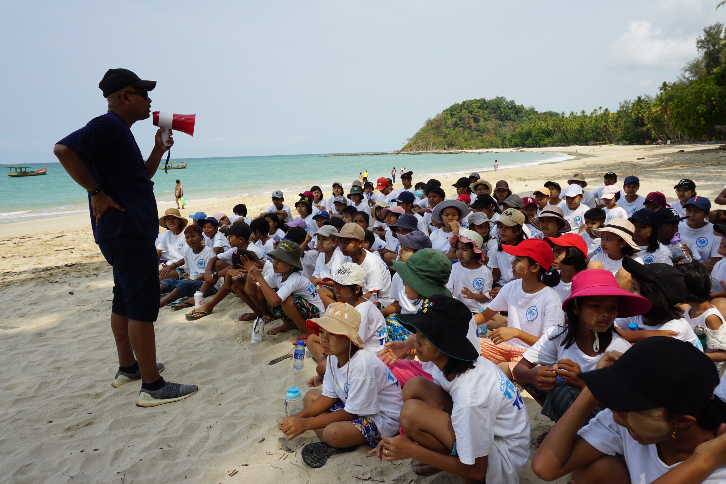 A man with a megaphone speaking to a group of children sitting on a beach with a hill and palm trees in the background.