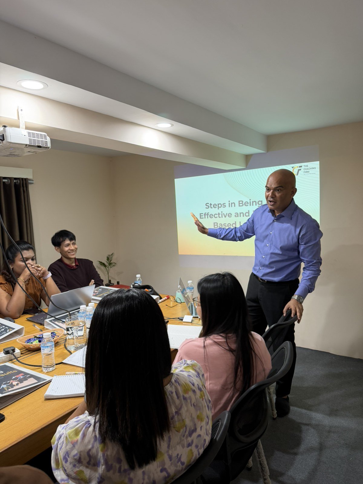 A professional man giving a presentation to a group of colleagues seated around a conference table in a meeting room, with a slide titled 'Steps in Being Effective and Based' projected on the wall.
