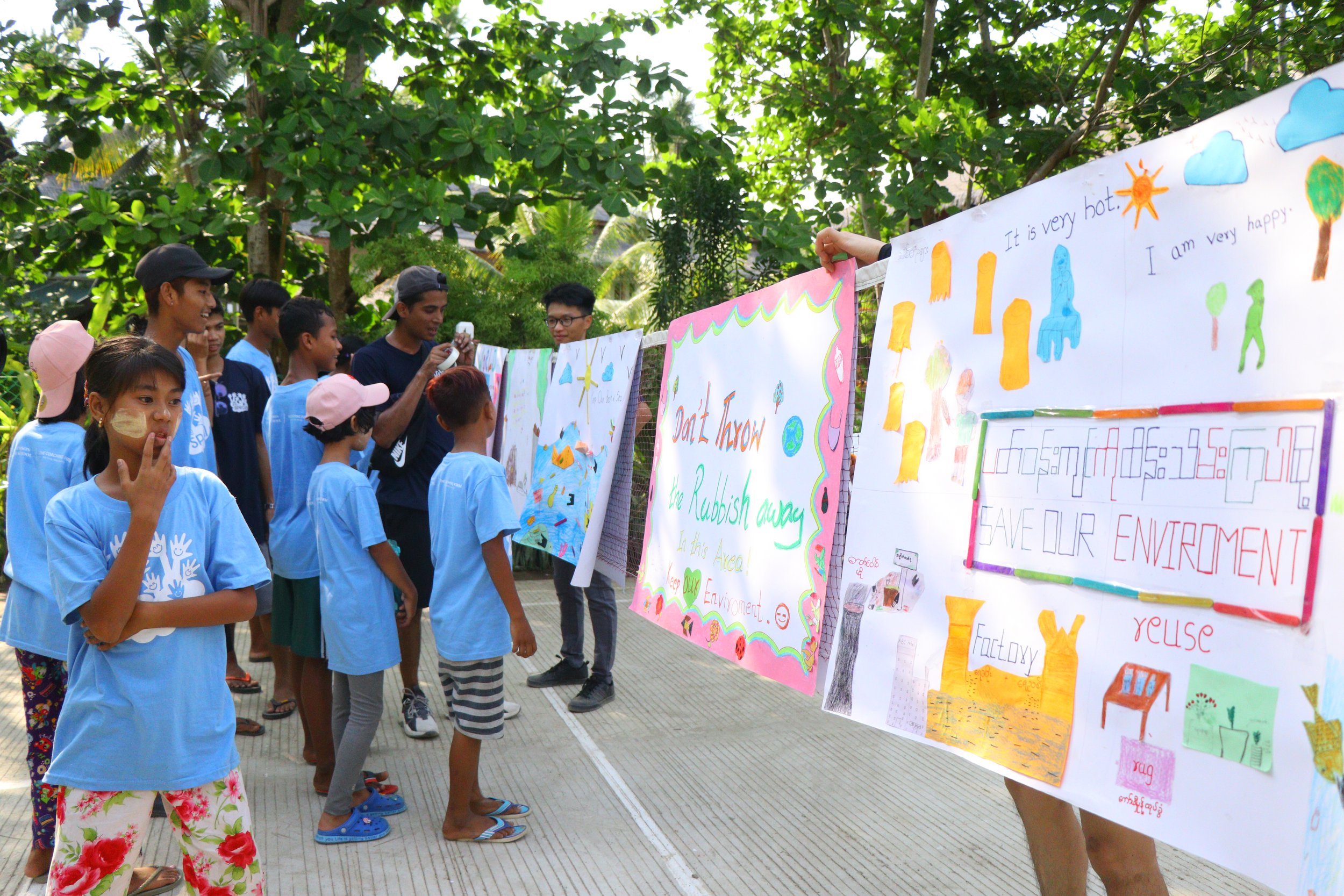 A group of children and teenagers in blue shirts are gathered outside, looking at colorful posters about environmental awareness displayed on a string. They are amidst lush green trees.