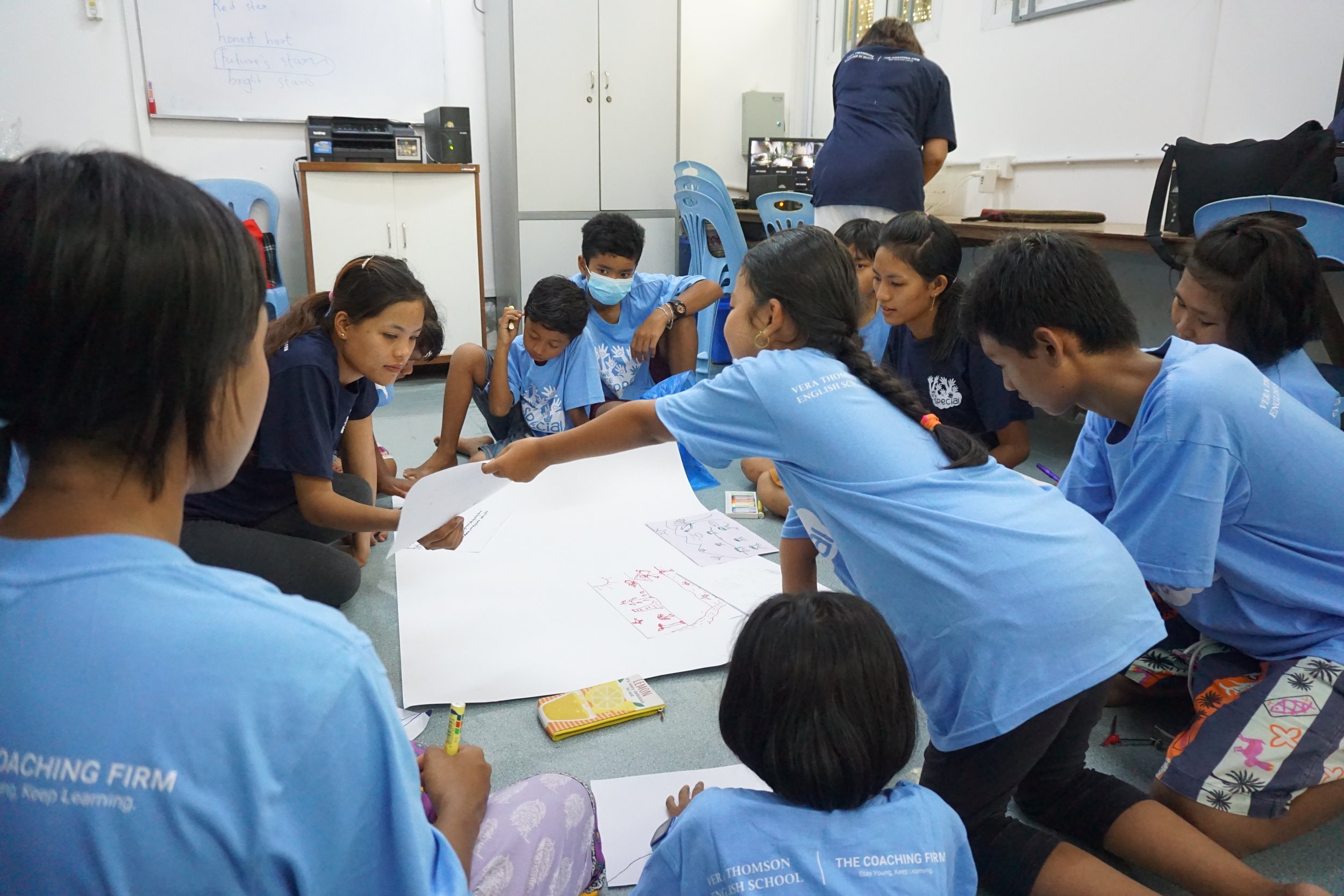 Group of children sitting on the floor and around a white table, working on drawings or writing, in a classroom with white walls and whiteboard. One child is wearing a face mask.