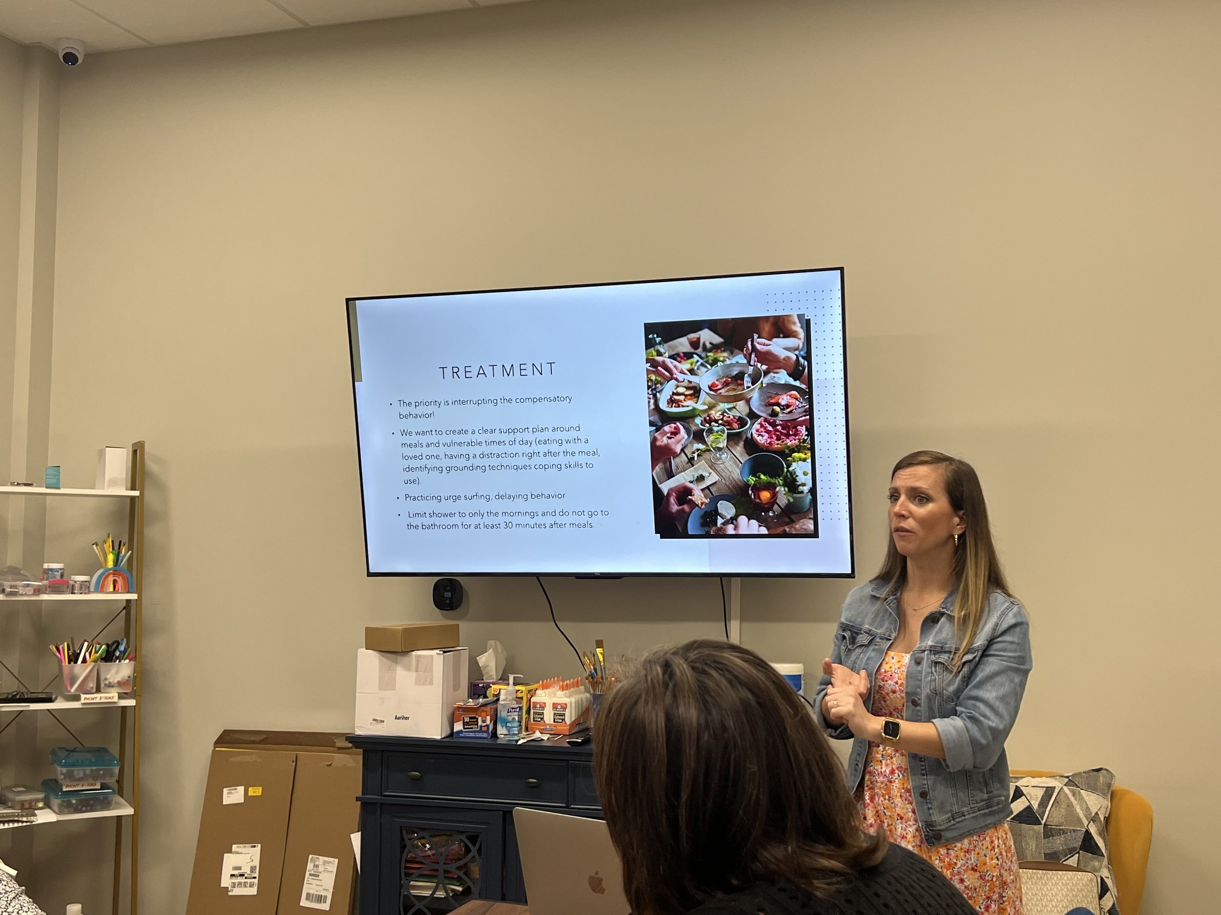 A woman with long hair, wearing a denim jacket, is standing in front of a group during a presentation. There is a large screen behind her displaying a slide about treatment, featuring a photo of various dishes with people serving themselves. The room has office supplies on shelves and a table with boxes and supplies.
