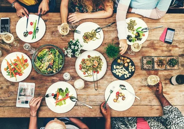 People sitting around a wooden dining table with various salads, drinks, and a smartphone, viewed from above.