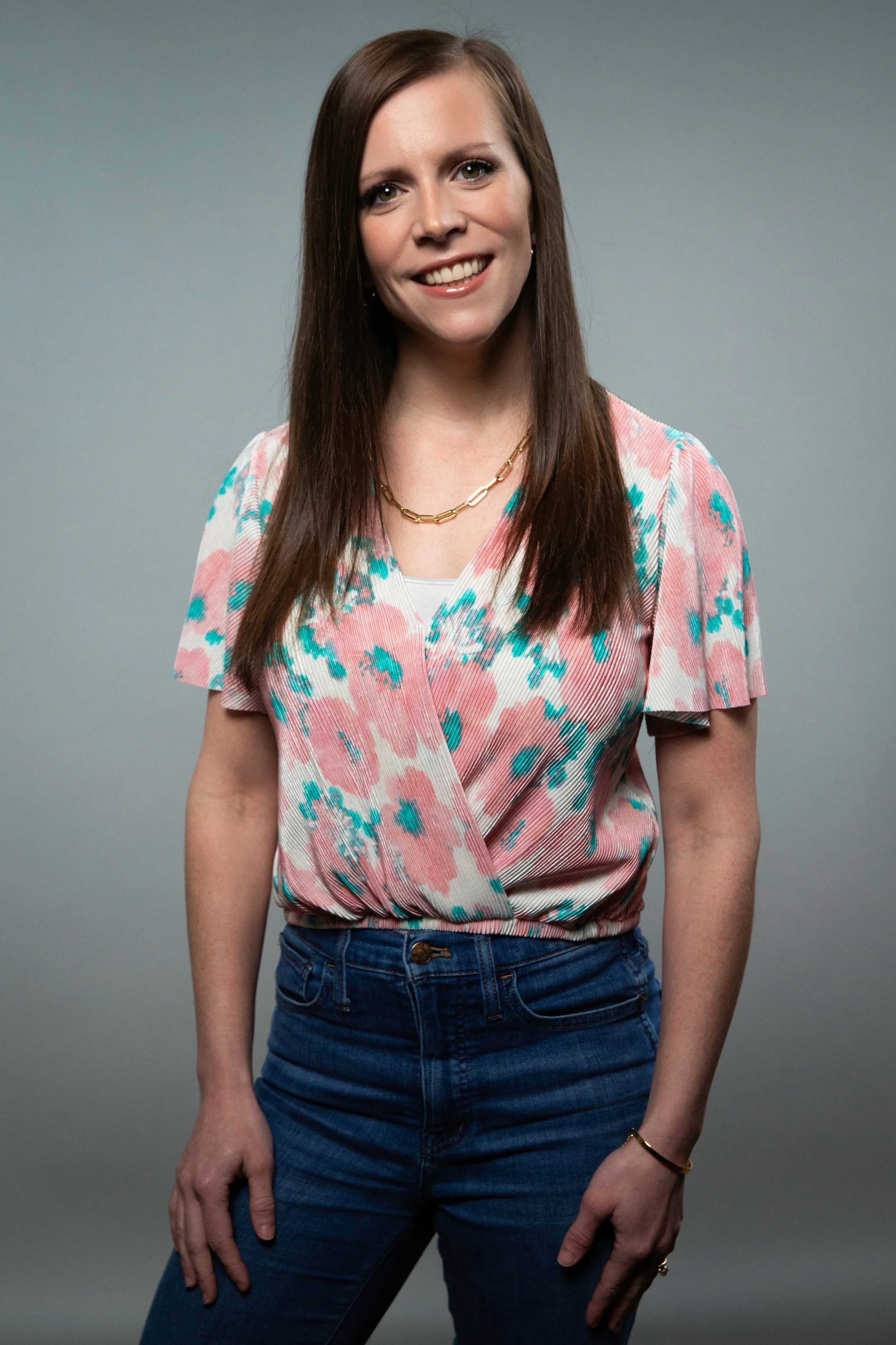A woman with long brown hair smiling, wearing a pastel pink and blue patterned top with a v-neck and short sleeves, paired with high-waisted blue jeans, against a plain gray background.