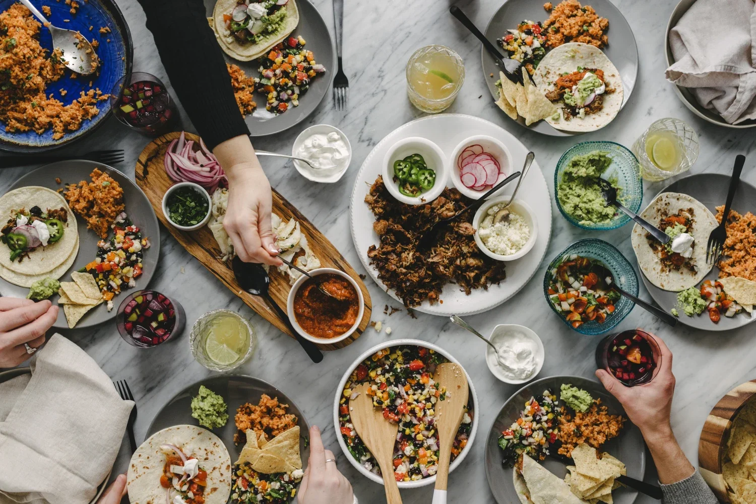 A table filled with Mexican dishes including tacos, rice, salsa, guacamole, and various side dishes, with glasses of lime water and people serving and eating.