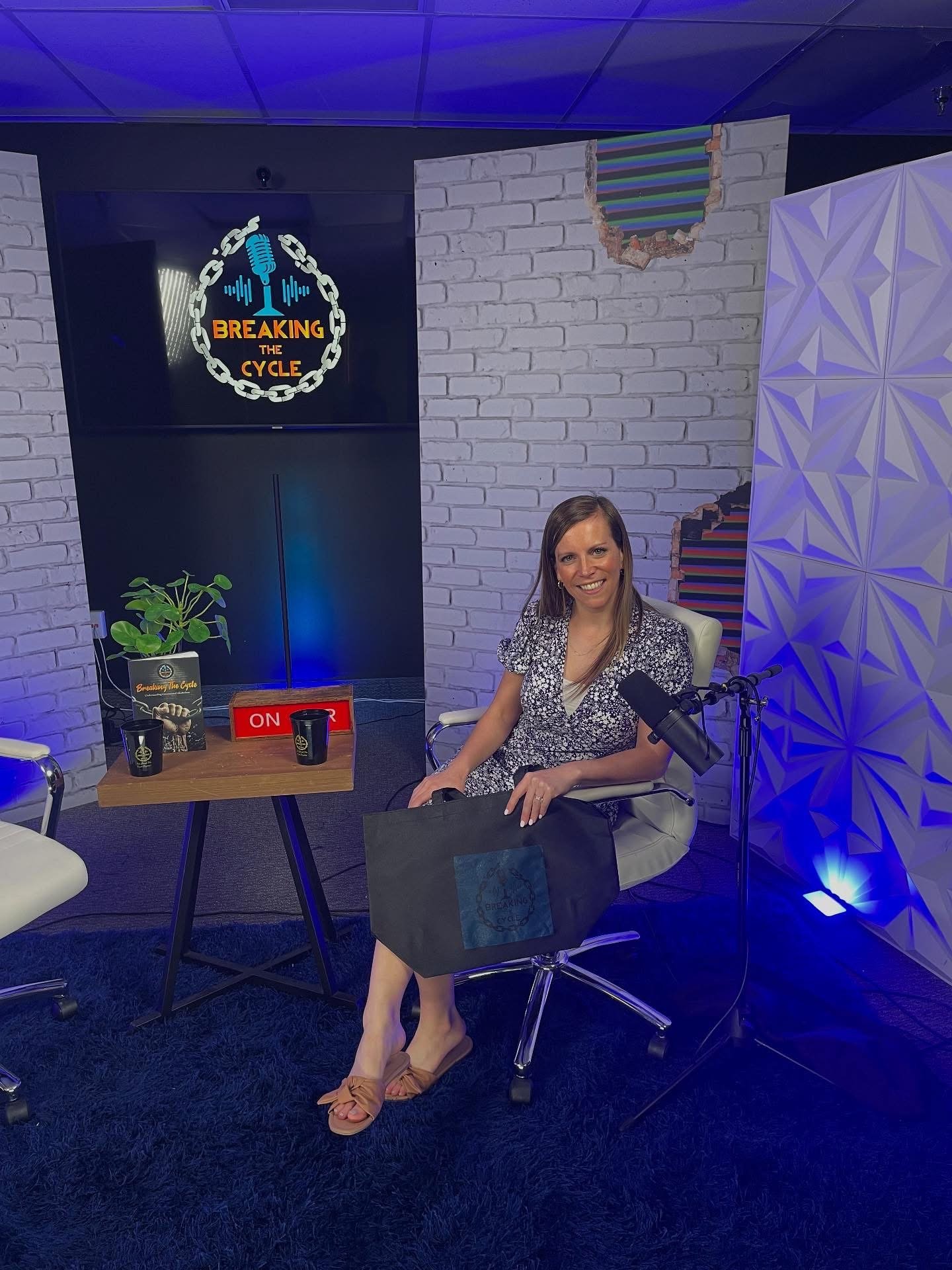 A woman sitting on an office chair in a room decorated with white brick walls, a sign that reads "Breaking the Cycle", a microphone, and a black tote bag. There is a table with two cups, a plant, and a book on it. The woman is smiling and wearing a floral dress, beige sandals, and has a microphone in front of her.