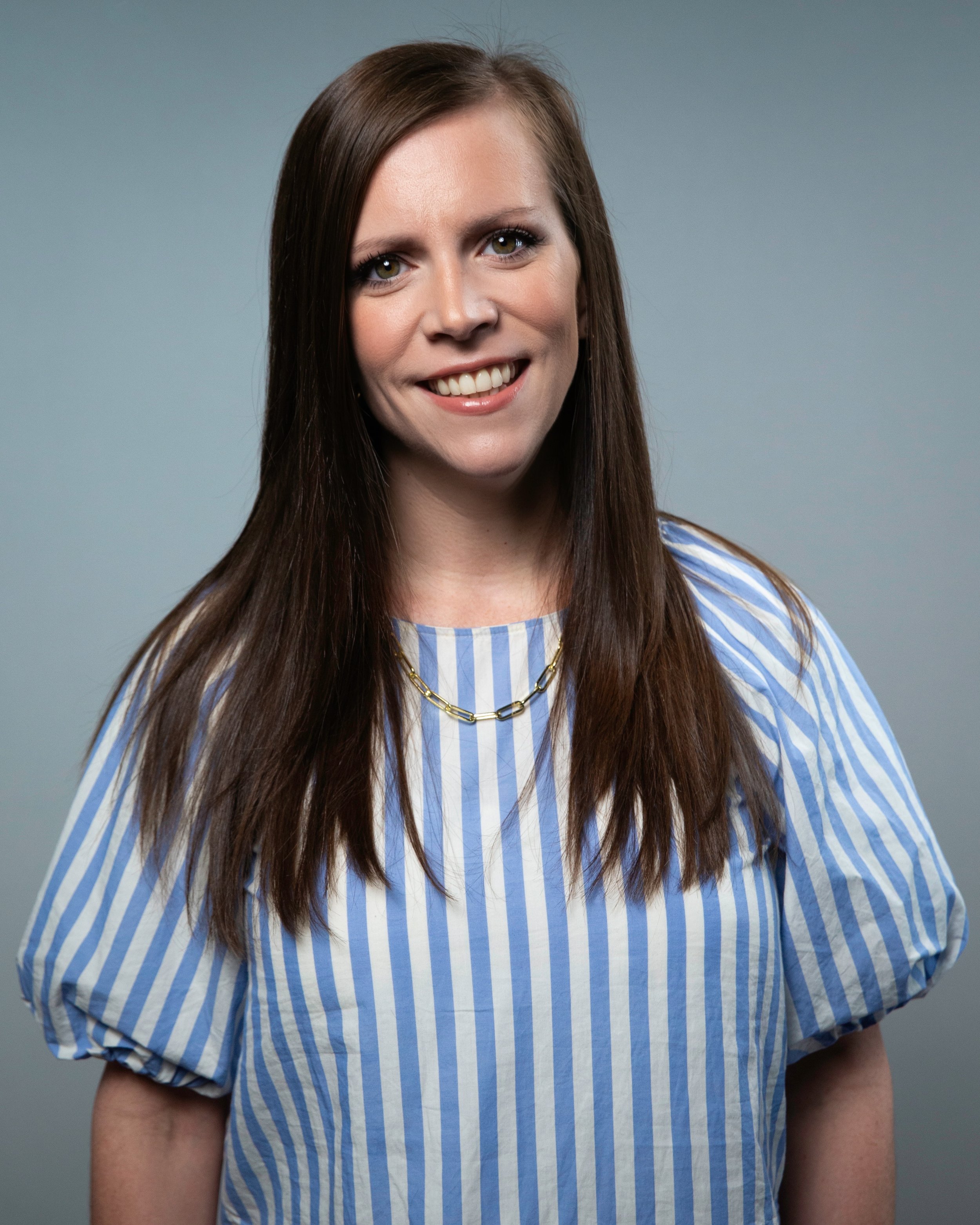 Close-up portrait of a woman with long brown hair, wearing a blue and white striped top and gold chain necklace, smiling against a gray background.