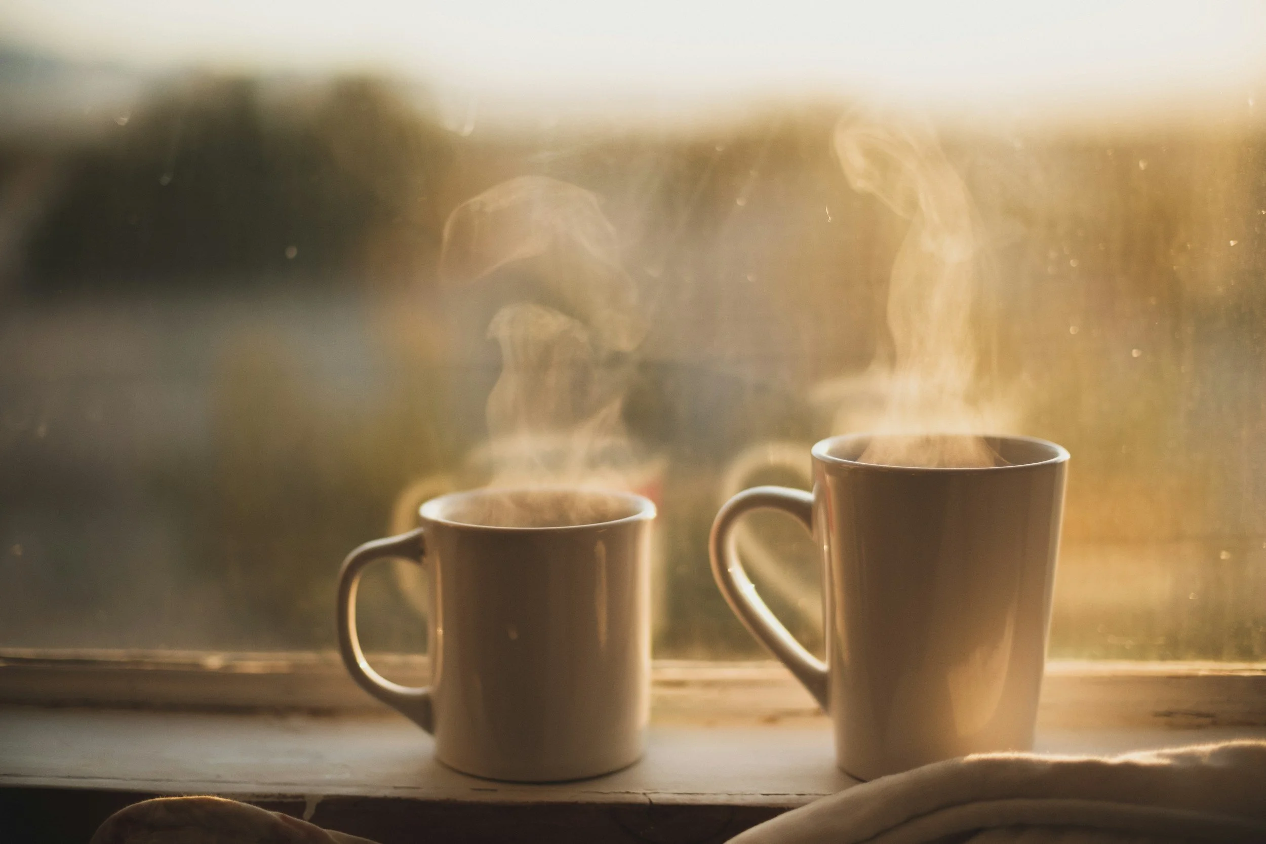 Two ceramic coffee mugs placed on a windowsill with steam rising from the hot beverages. Sunlight illuminates the steam and creates a warm, cosy scene.