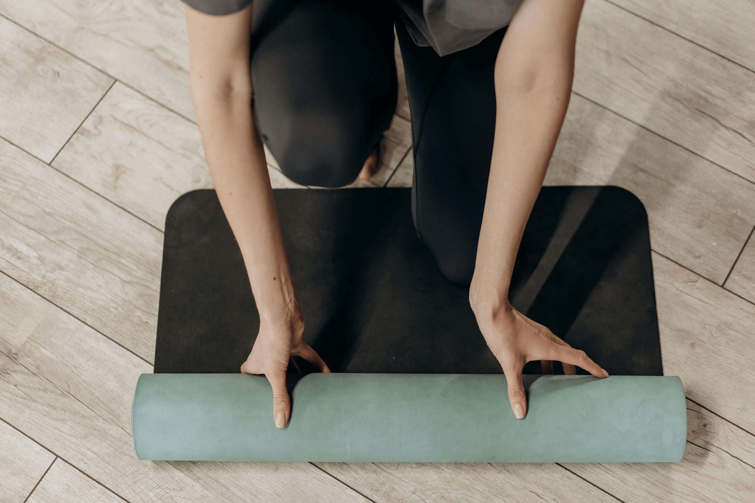 Top view of a person kneeling on a black yoga mat and unrolling a mint-green yoga mat on a wood floor