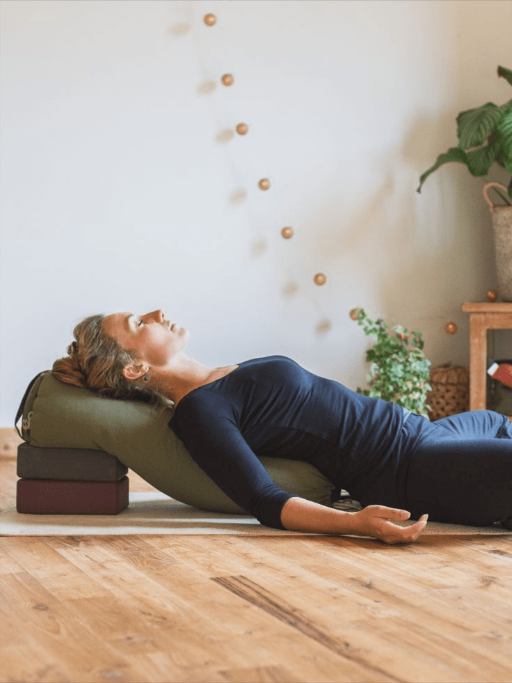 Woman lying in a supported restorative yoga pose on a mat in a bright room