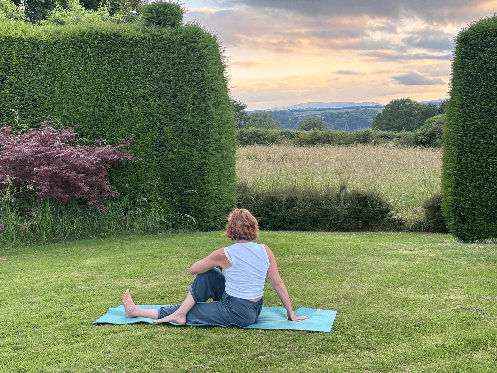 Woman stretching on a yoga mat in a garden with landscaped hedges and a countryside sunset view
