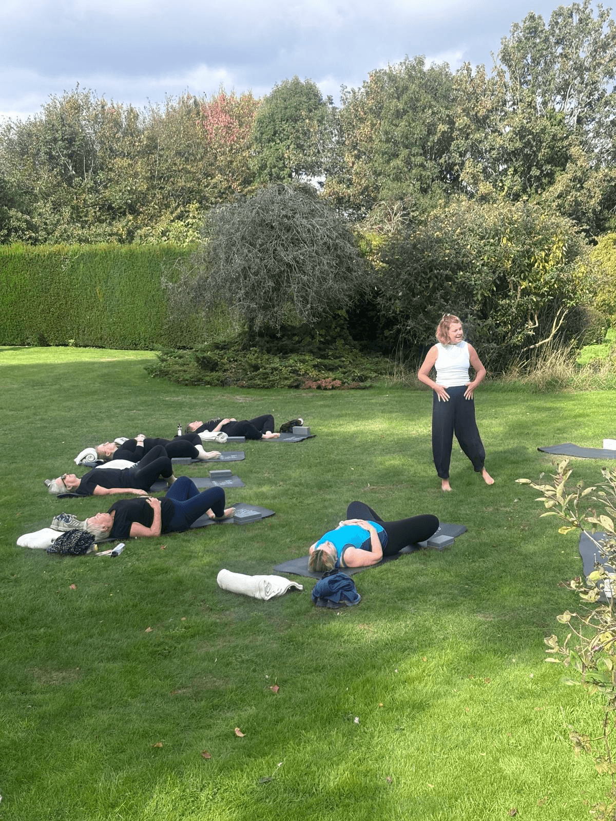 Outdoor Pilates class with participants exercising on mats in a garden while an instructor stands nearby.