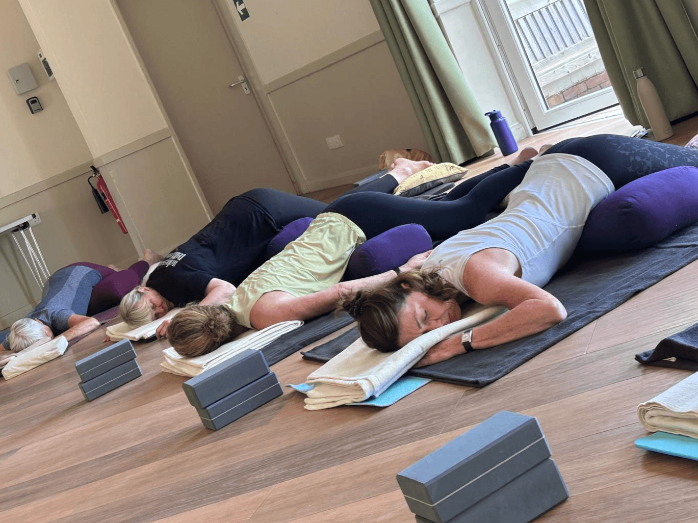 Group of people lying on yoga mats during a restorative yoga class with bolsters and folded towels.