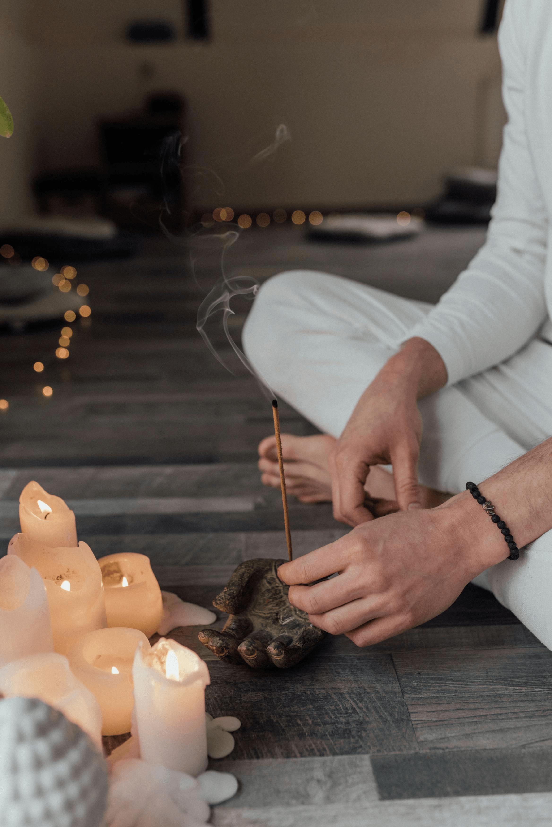 Person in white clothing performing a candlelit ritual with incense over a dark wooden table.