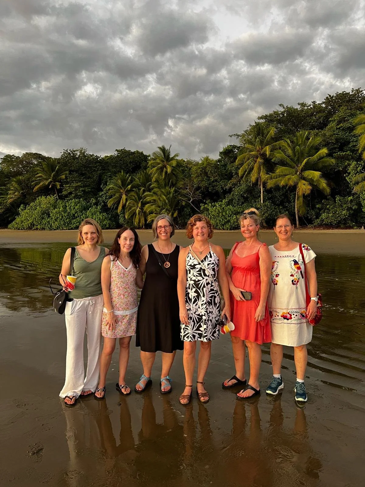 Six women standing barefoot on a tropical beach with palm trees and storm clouds.