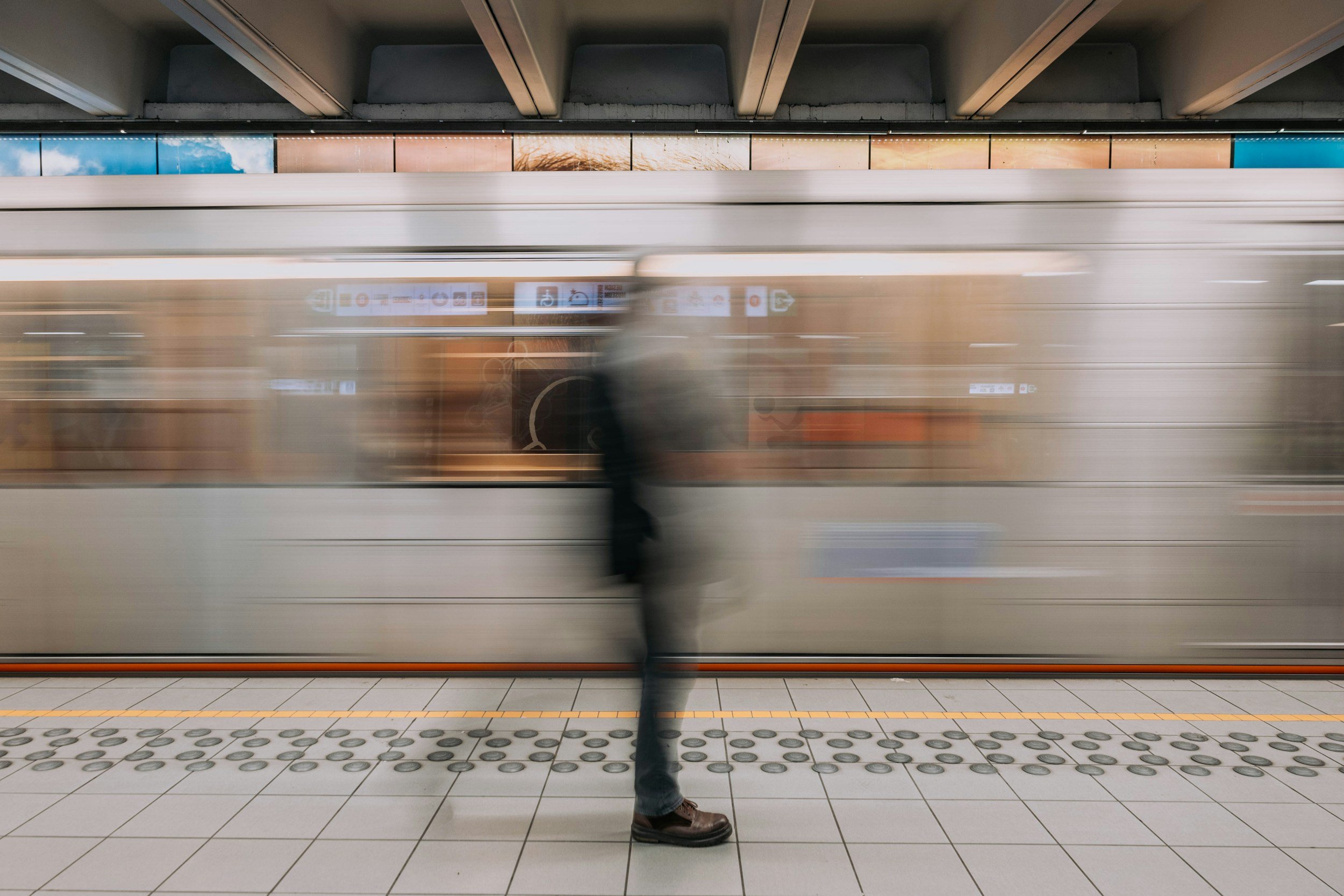 Subway train at a station with a person standing on the platform, blurred motion of the train passing by, and part of the platform with tactile paving.