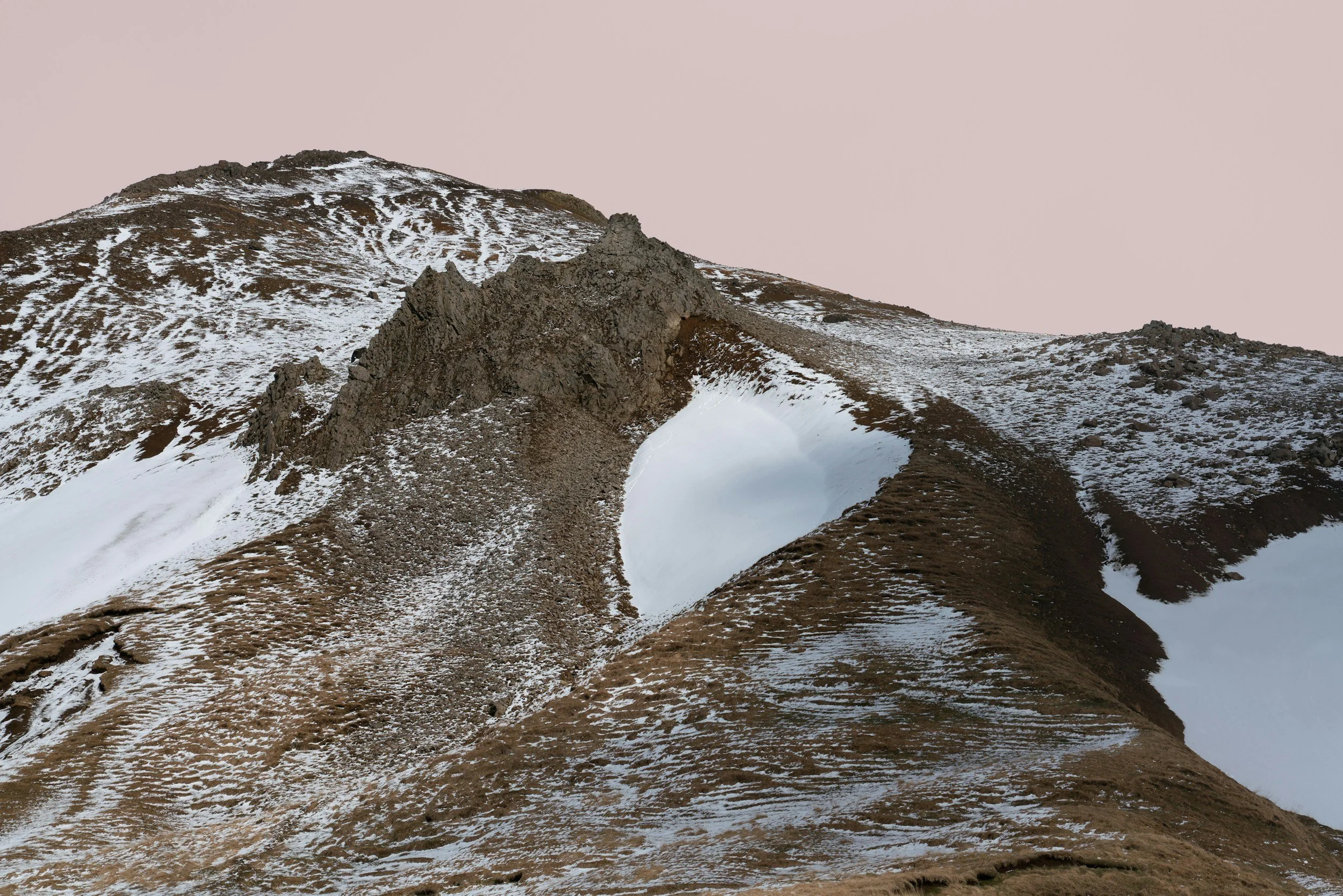Snow-capped mountain with rugged rocky outcrop, brown and white patches of snow, and a pastel pink sky in the background.