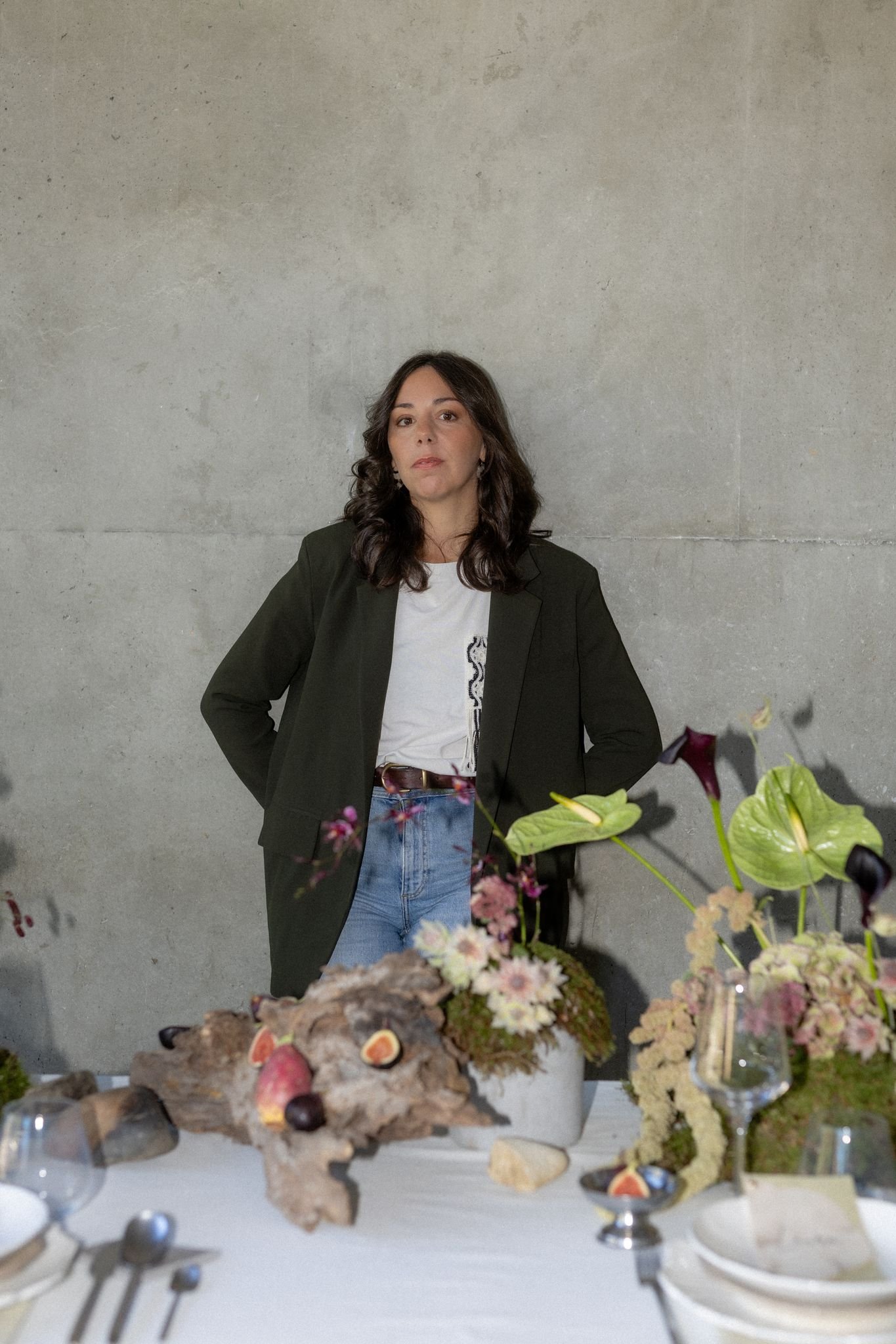Une femme regarde l'appareil photo, debout devant un mur en béton visible en arrière-plan. Elle porte un blazer noir, un t-shirt blanc avec un motif, et un jean. Des fleurs et des objets de décoration sont visibles sur une table à l'avant de l'image.