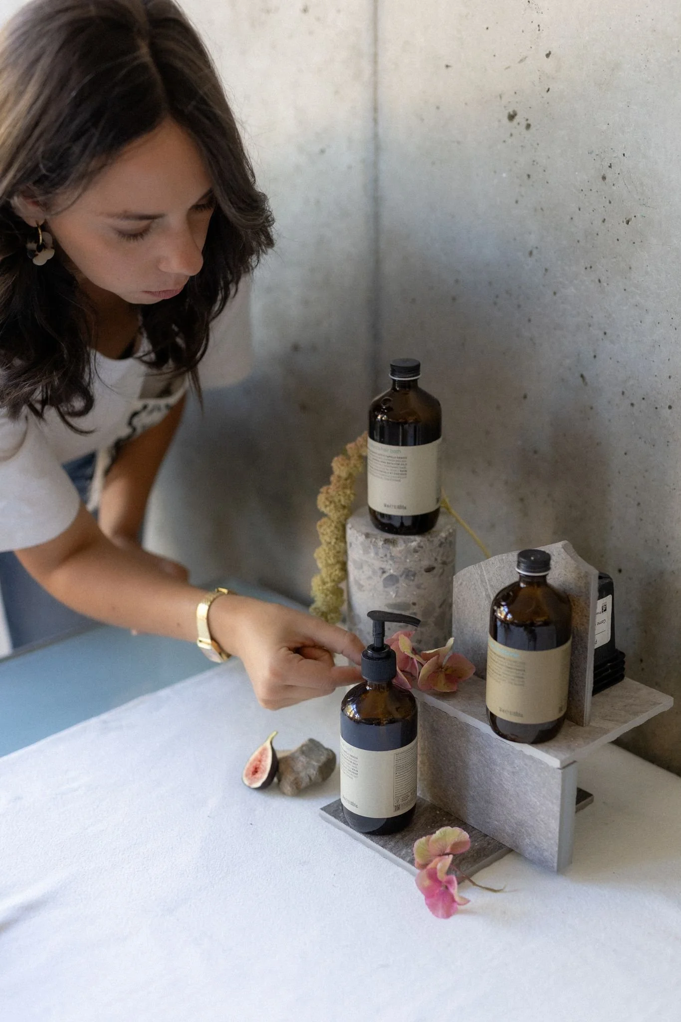 Une femme avec des cheveux bruns foncés, portant des boucles d'oreilles blanches, utilise un distributeur de savon dans un environnement intérieur minimaliste avec un mur en béton.