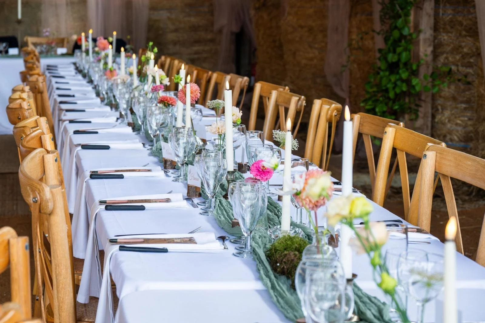 Long banquet table decorated with candles, flowers, glasses, and napkins in a rustic setting.