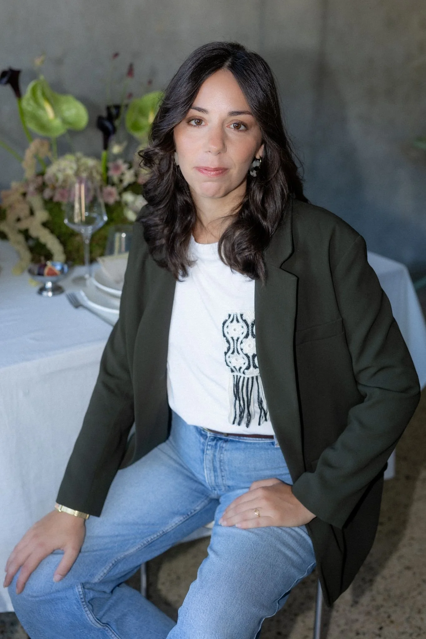 Femme assise portant un blazer noir, une chemise blanche avec un motif noir, et un jean, assise à une table avec un décor floral en arrière-plan.