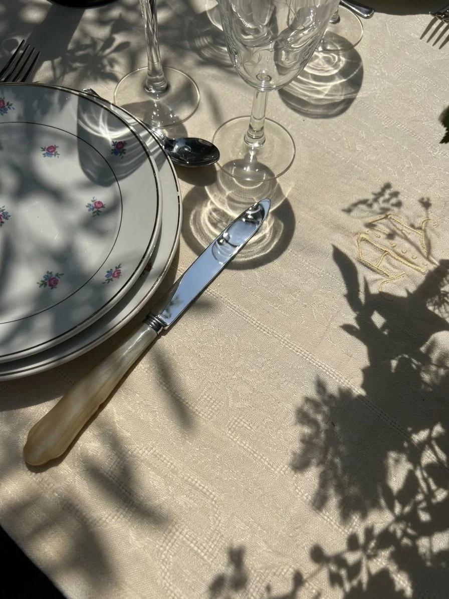 Une table élégante avec des assiettes en porcelaine blanche ornées de petites fleurs, un couteau avec un manche en matière de nacre, deux verres à vin vides, et des ombres de plantes sur une nappe beige, sous la lumière du soleil.