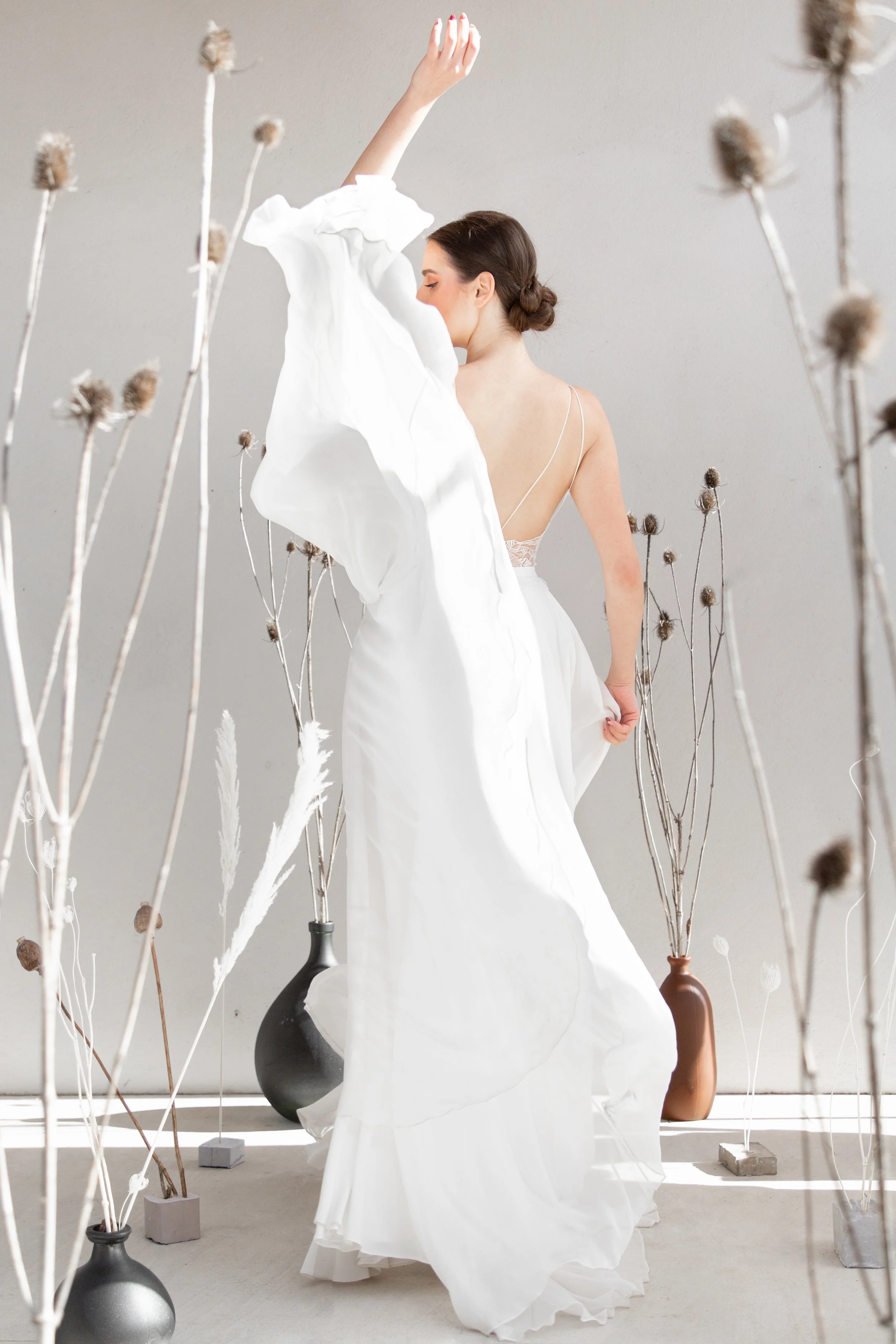 Une femme en robe blanche posant dans une pièce sombre, entourée de vases avec des branches séchées.