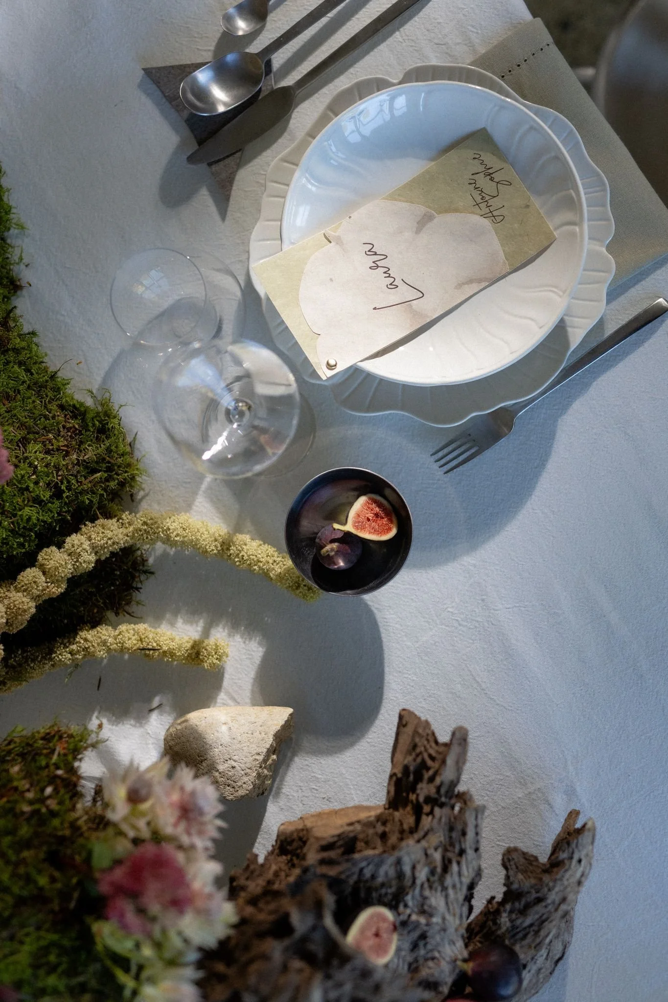 Une table décorée avec une assiette blanche, un couteau, deux cuillères, un verre à vin, un bol avec des figues, et un menu avec l'inscription "Love" en français. Décoration florale en premier plan.