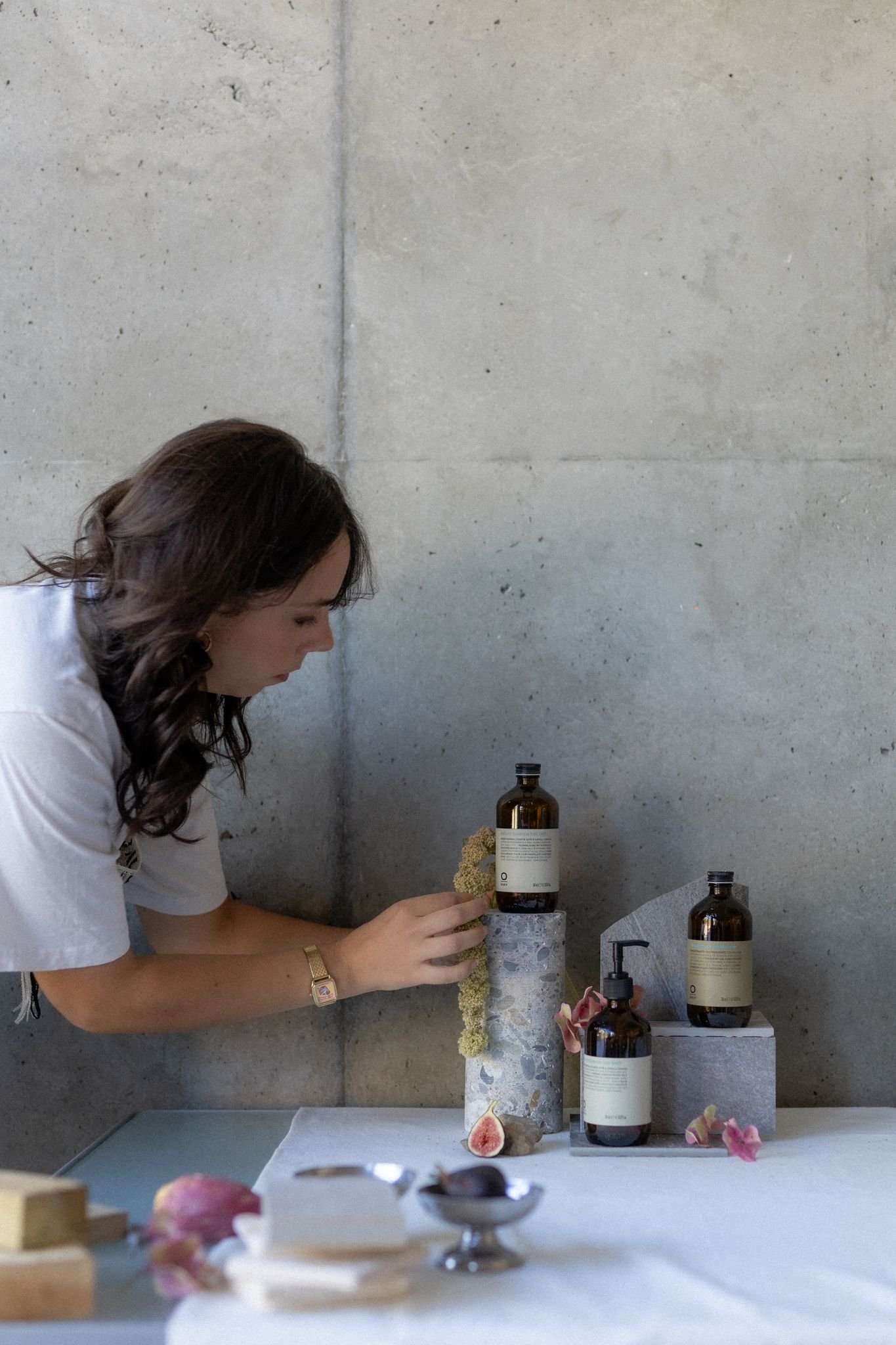 Une femme approche une bouteille de produits de beauté ou de parfum sur une table blanche devant un mur en béton, avec des fleurs et un figuier coupé pour la décoration.