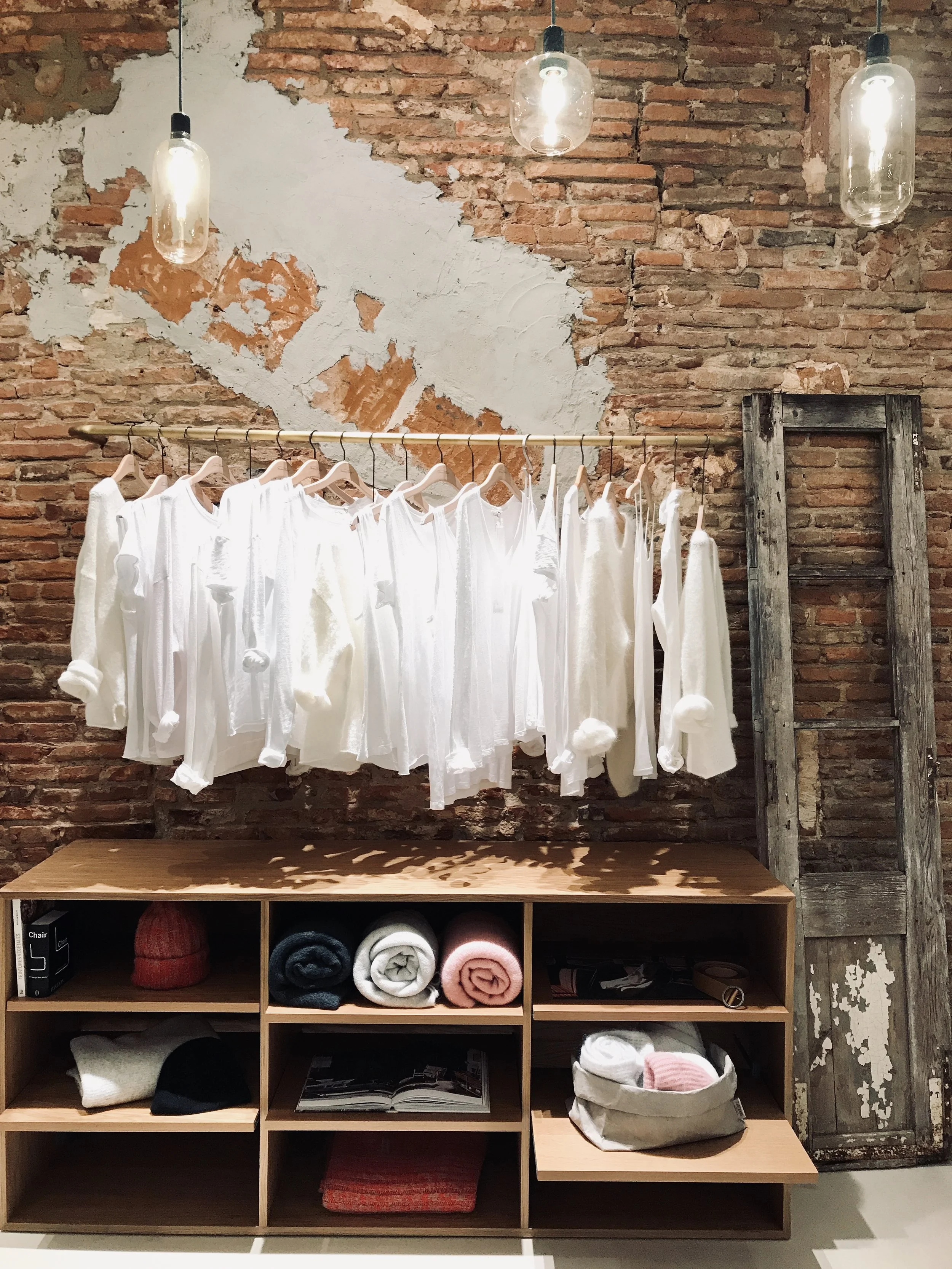 Vêtements de différentes couleurs suspendus sur une tige en or au mur, devant un mur en briques avec une peinture partielle. Une mannequin porte une veste en jean et une robe grise. Une étagère en bois contient des vêtements pliés, un livre et des accessoires.