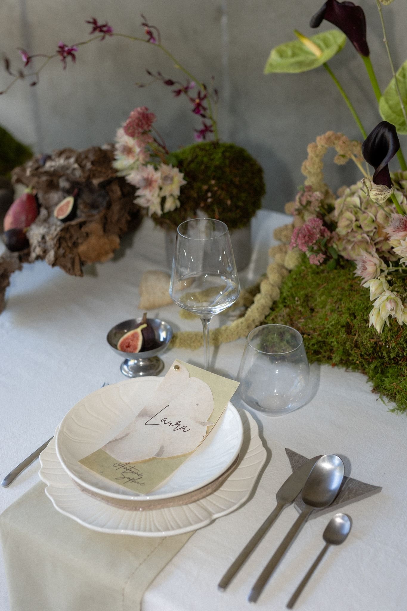 Décoration de table élégante avec un plateau blanc, une branche d'arbre en papier avec une note au nom de Laura, un verre à vin, et des ustensiles en métal argenté sur une nappe blanche, avec des fleurs et des éléments végétaux en arrière-plan.