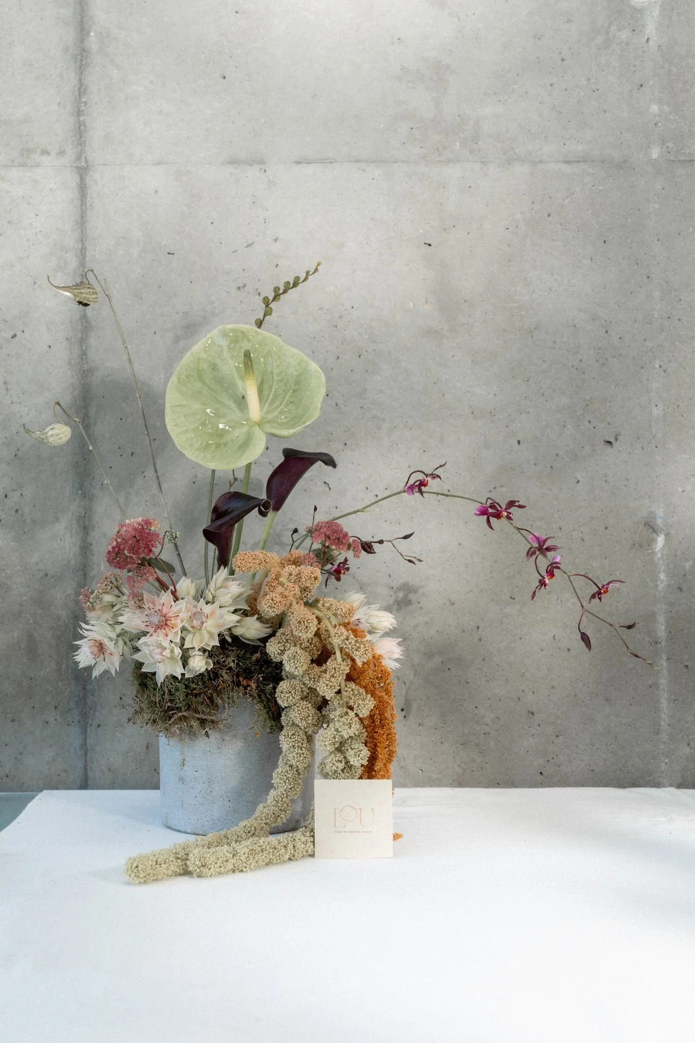 Un arrangement floral dans un vase gris clair, comprenant diverses fleurs et plantes, avec un fond en béton.