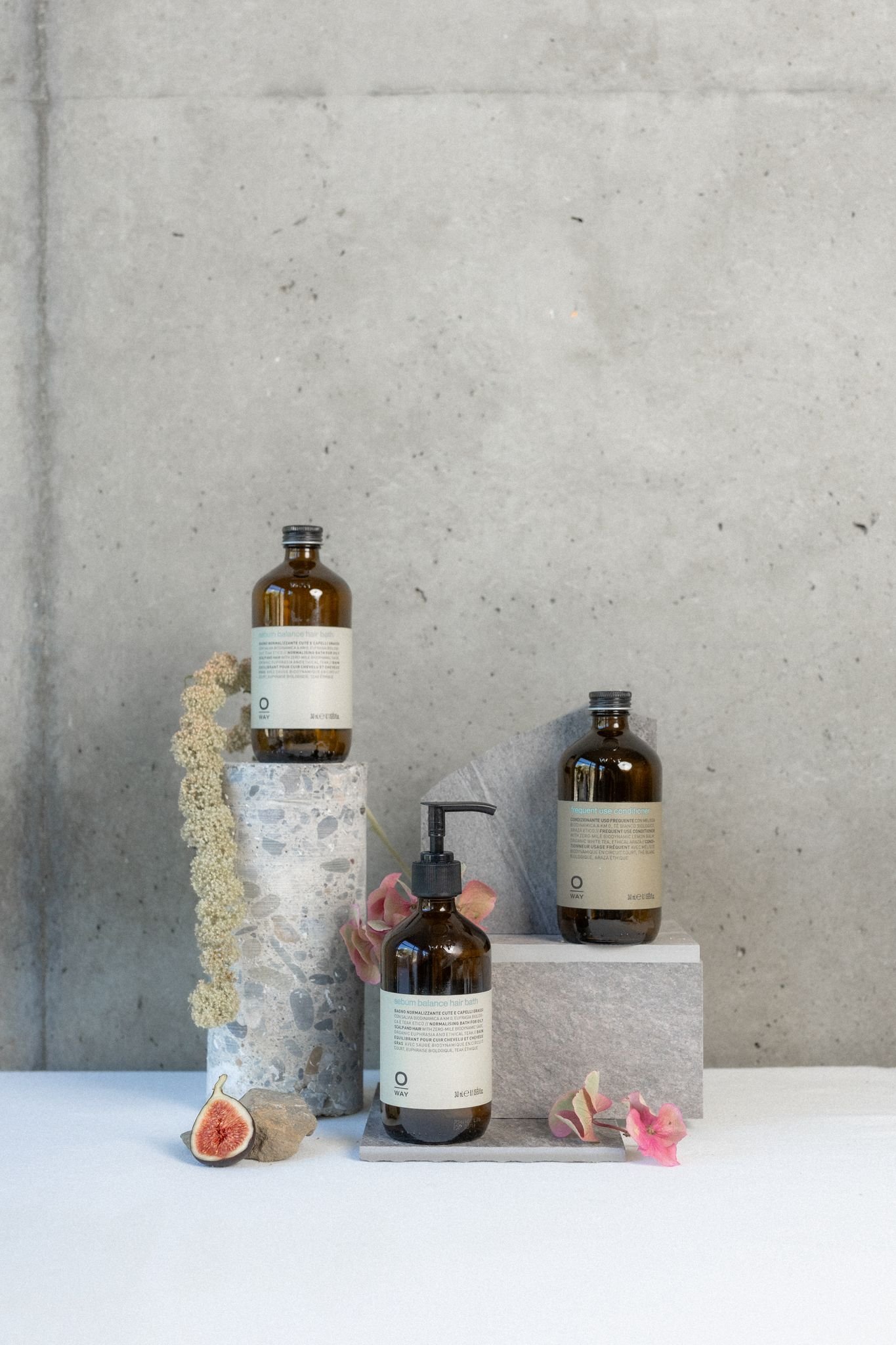 Bouteilles de produits cosmétiques sur des piédestals en pierre dans un décor minimaliste avec des fleurs et une figue ouverte.