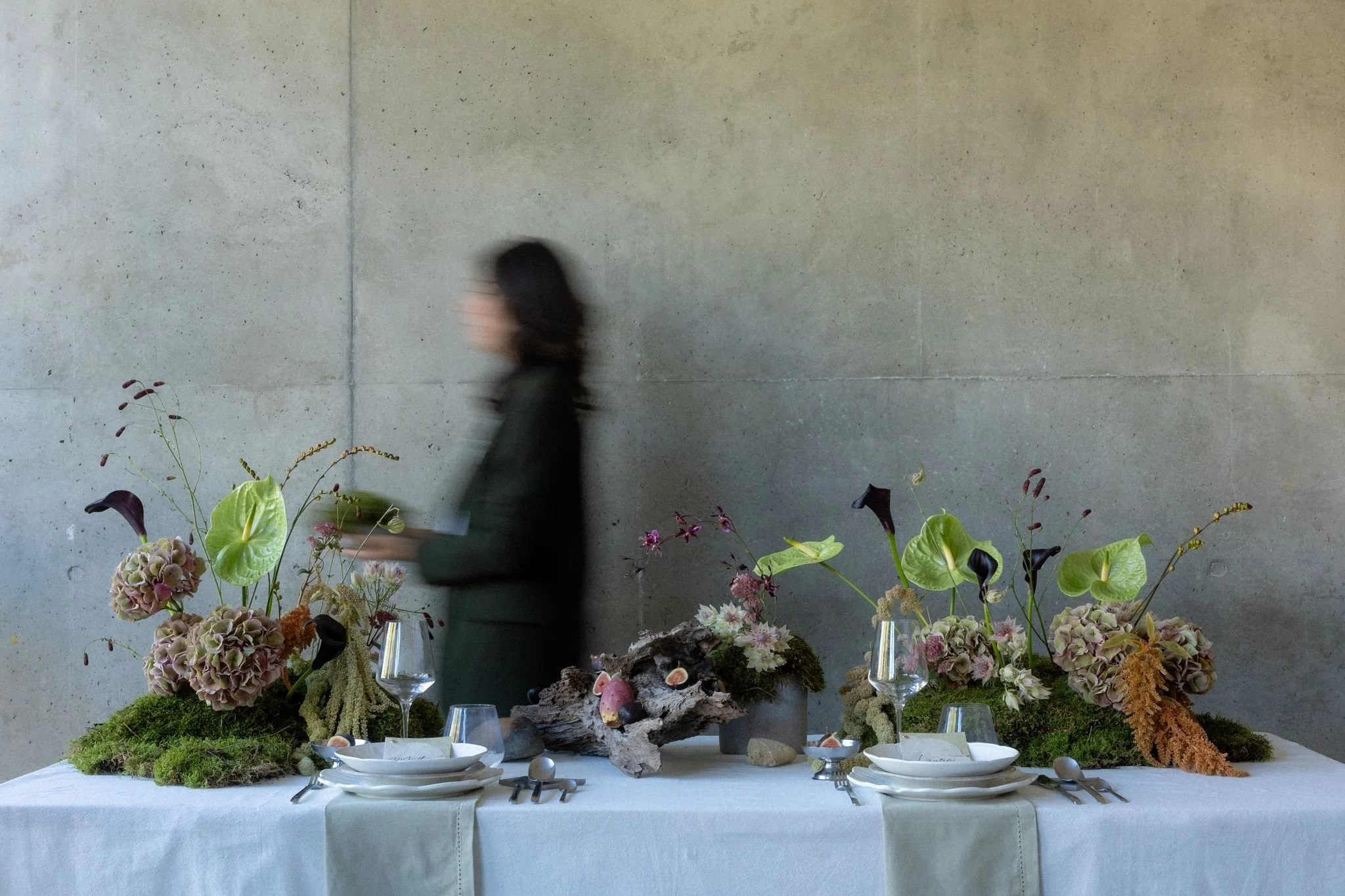 Une table dressée avec un arrangement floral au centre, comprenant des hortensias, des callas, et autres fleurs, sur un tapis de mousse, sous un mur en béton. Des assiettes, couverts, verres et petits plats sont disposés autour de l'arrangement.