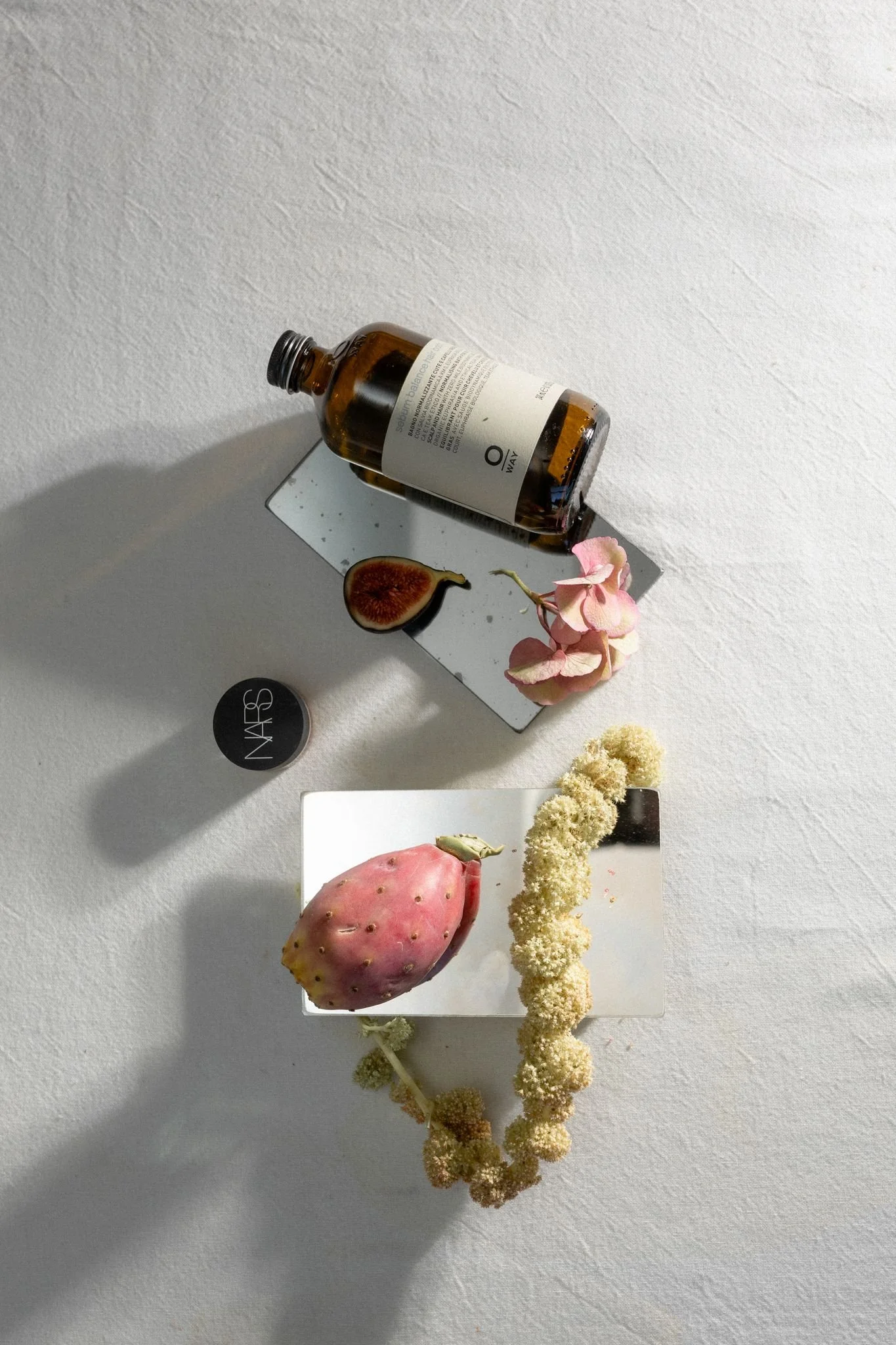 Une bouteille en verre marron à moitié remplie, une figue coupée en deux, un figuier de Barbarie, un petit bouquet de fleurs roses, un bouquet de fleurs blanches, et un fond blanc.