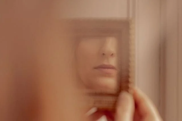 Une femme regardant dans un miroir, en partie obscurcie par une surface translucide ou une boîte en bois.