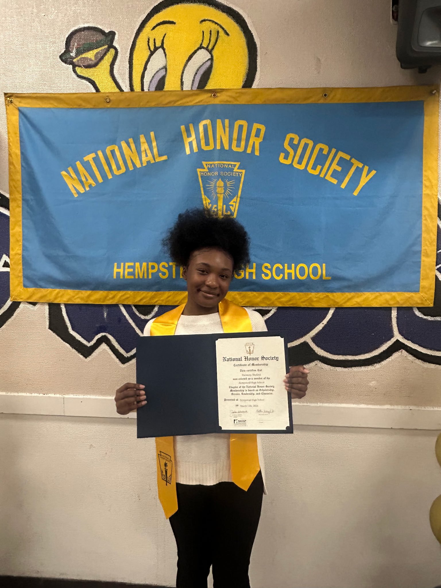 A person standing and holding a certificate, standing in front of a wall with a blue and yellow banner that reads 'National Honor Society.' The banner features a torch emblem, and a large cartoon-style yellow smiley face painted on the wall above the banner.