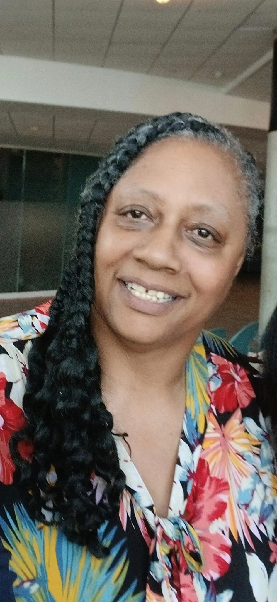 A woman with dark curly hair styled in a long braid, smiling, wearing a colorful floral shirt, in an indoor setting.