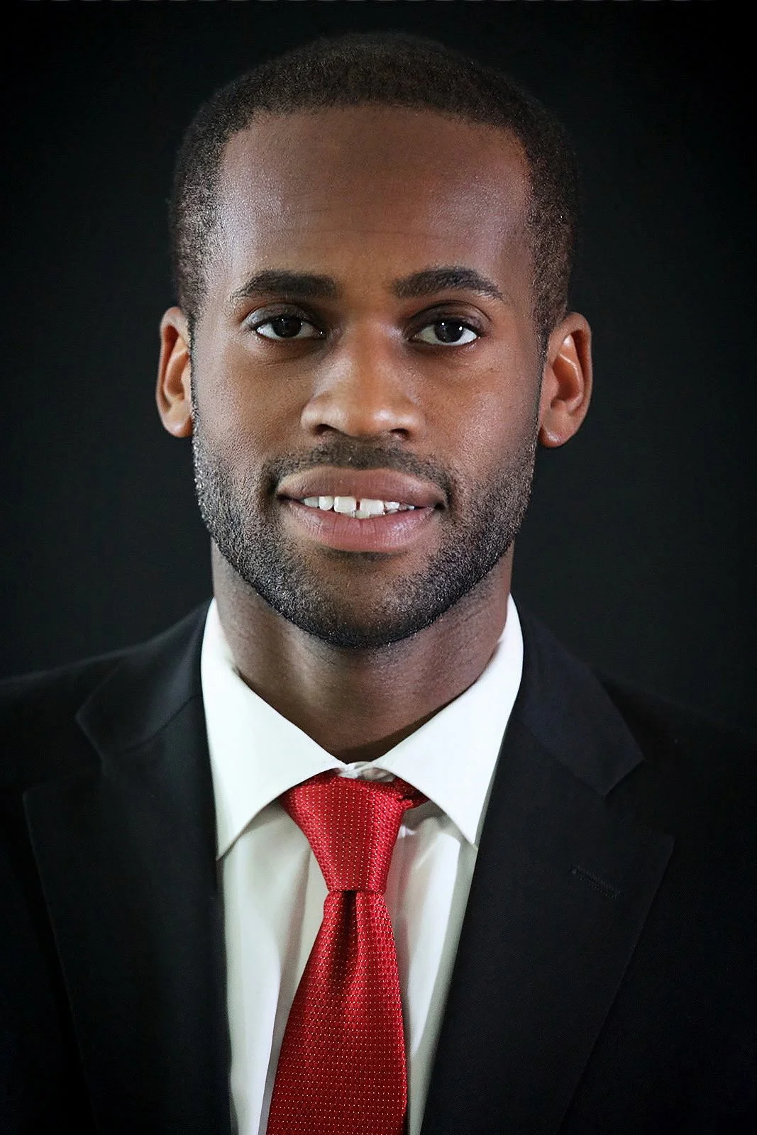 A portrait of a man wearing a black suit, white shirt, and red tie, with a dark background.