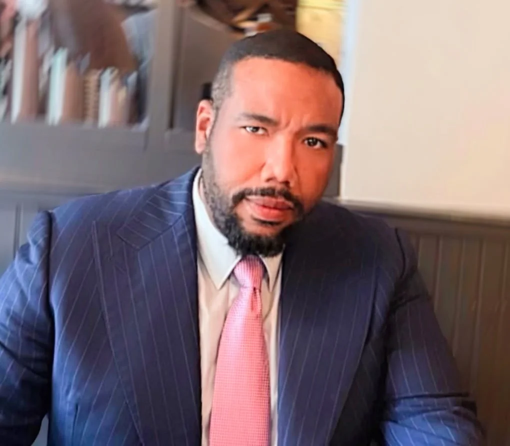 A man with a beard and short hair wearing a dark pinstripe suit, white shirt, and pink tie, sitting at a table in a restaurant or café.