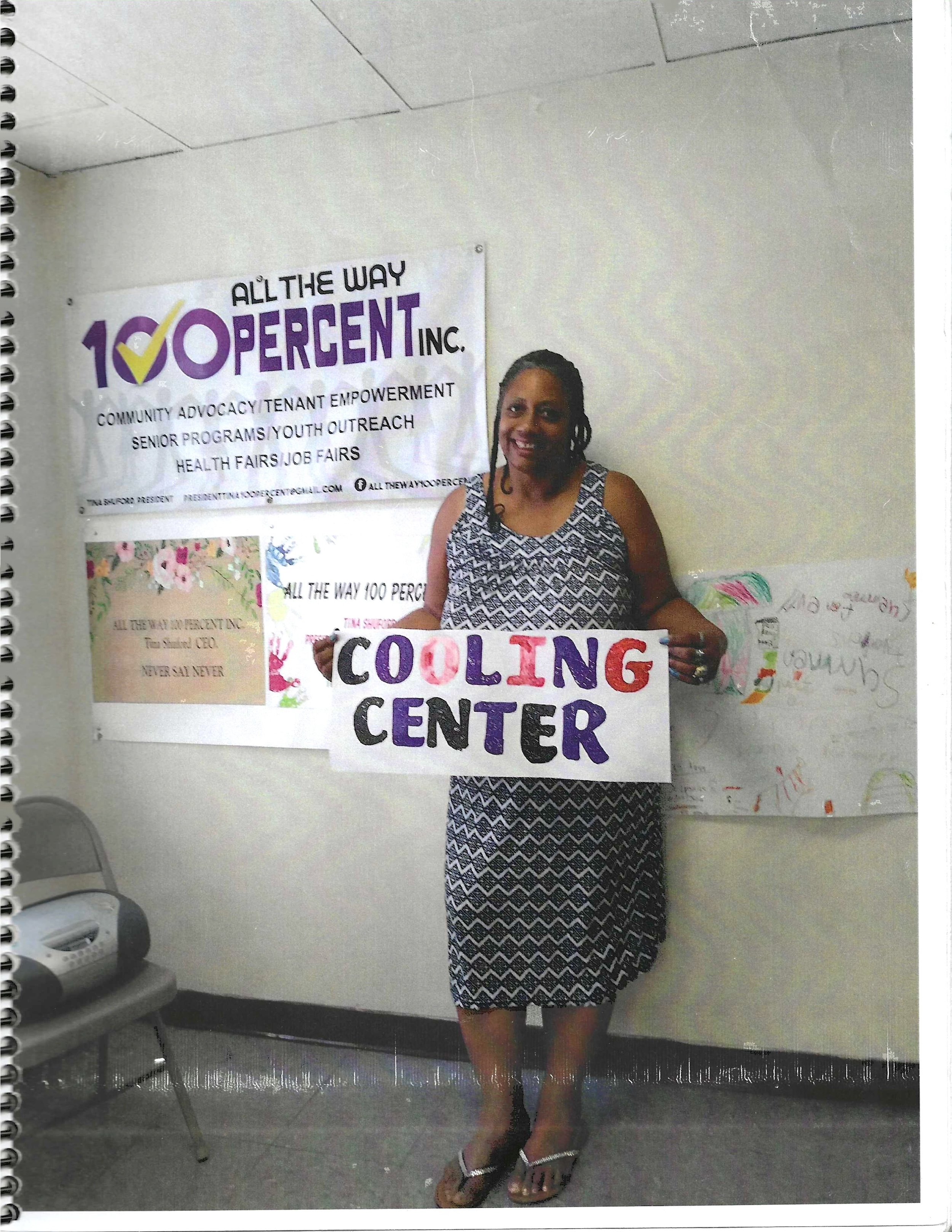 Woman standing in front of a wall with posters, holding a colorful sign that reads 'Cooling Center.' She is wearing a sleeveless, patterned dress and high-heeled sandals.