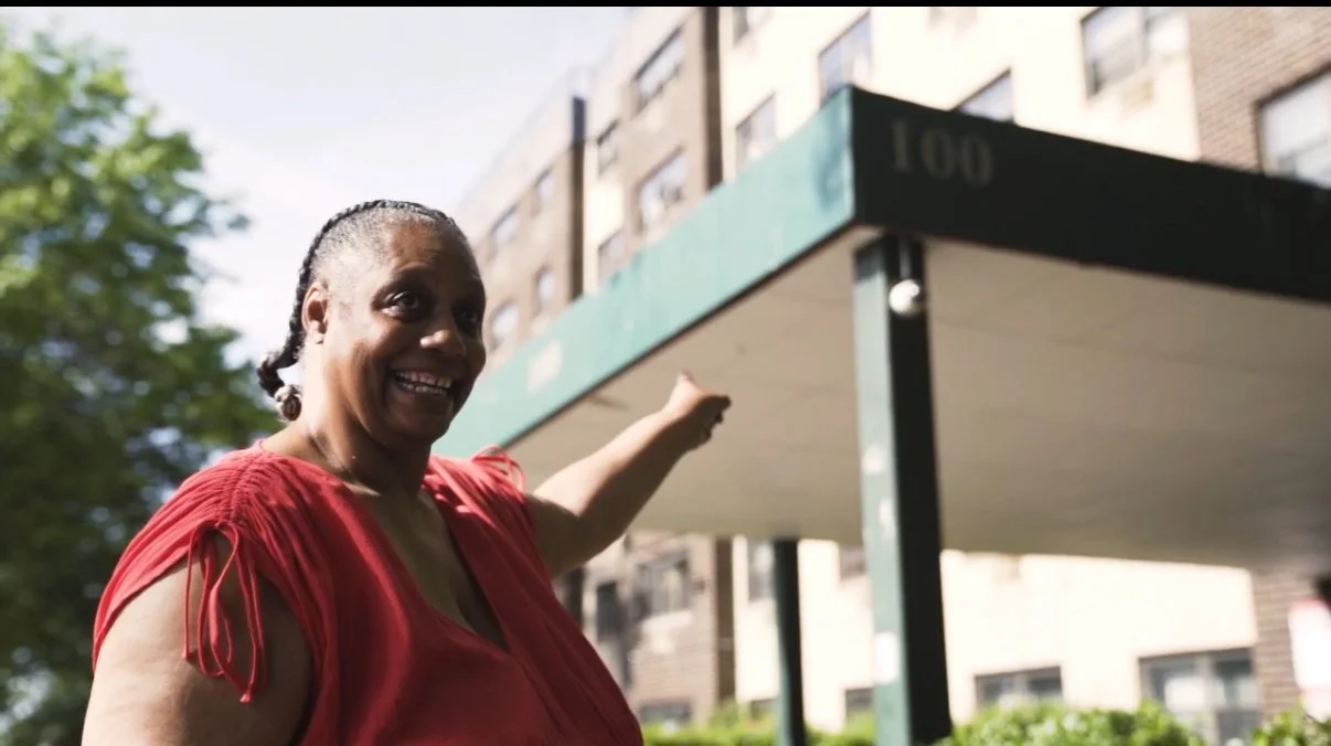 A woman in a red top smiling and pointing towards a building with the number 100 on it.