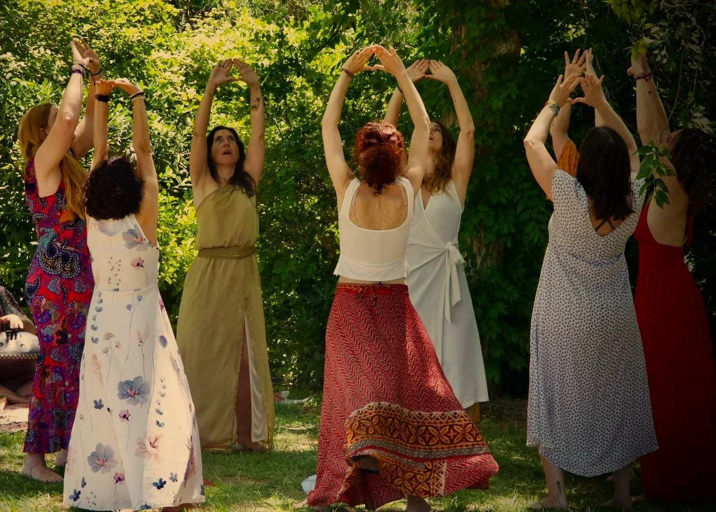 Varias mujeres de diferentes edades participando en una actividad al aire libre, con muchas plantas verdes y árboles en el fondo, levantando los brazos y formando triángulos con las manos.