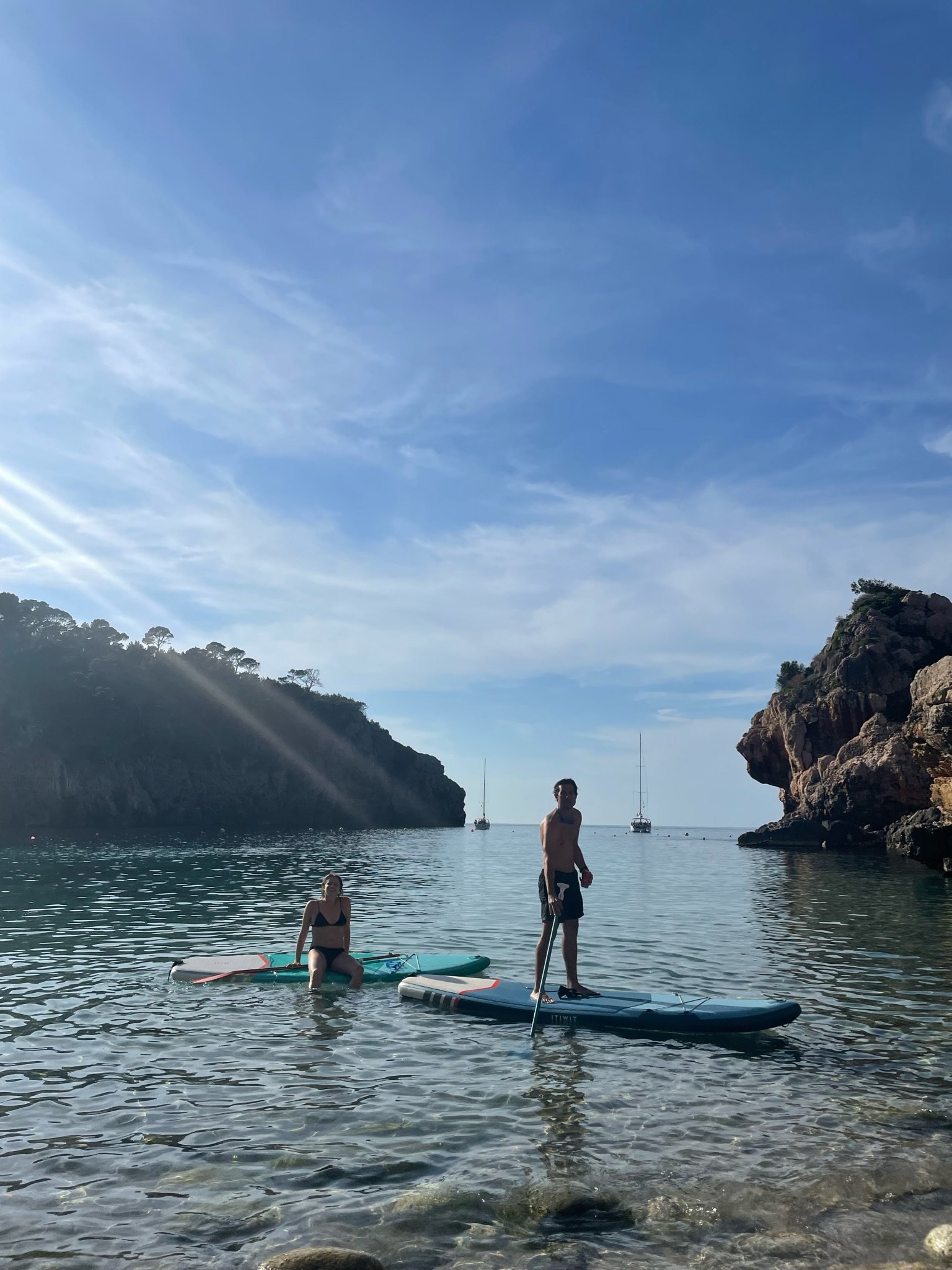 People paddleboarding on clear, calm water near rocky cliffs and green trees under a blue sky with wispy clouds.