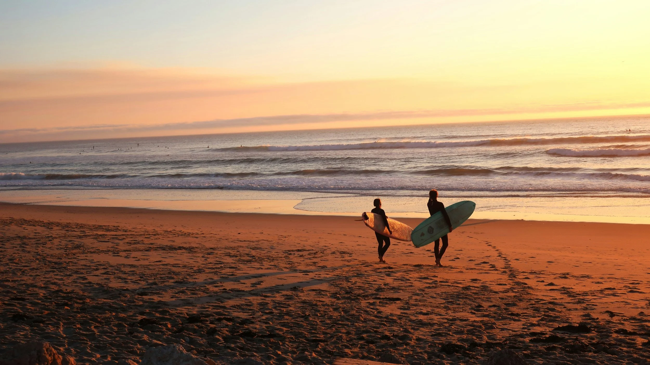 Two surfers walking on the beach at sunset, carrying surfboards, with waves and surfers in the ocean in the background.