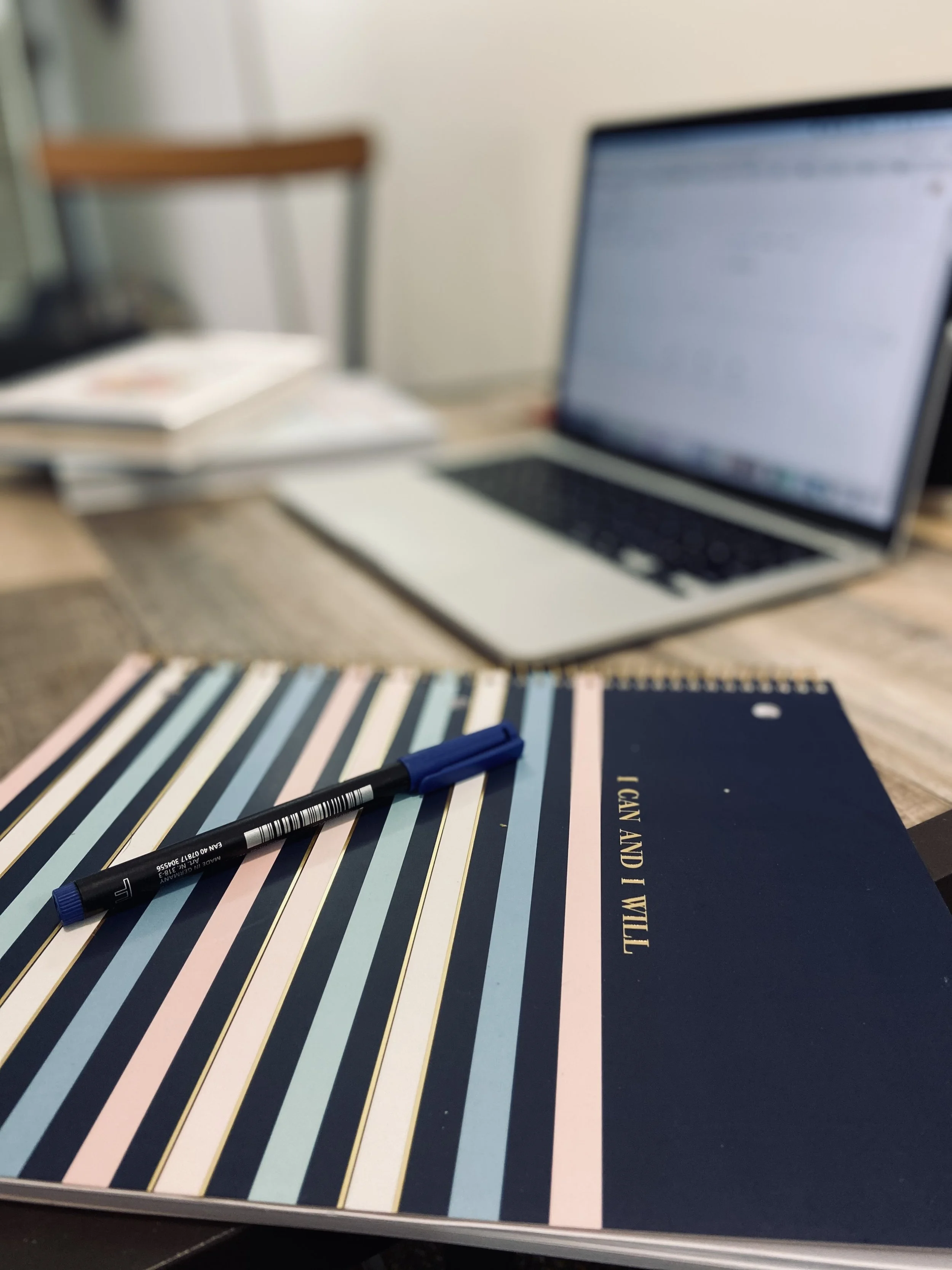 A closed navy blue notebook with gold lettering and a striped pattern on the cover, a blue pen resting on top, and a laptop with a blurred screen in the background on a wooden desk with an open book and a blurred chair.