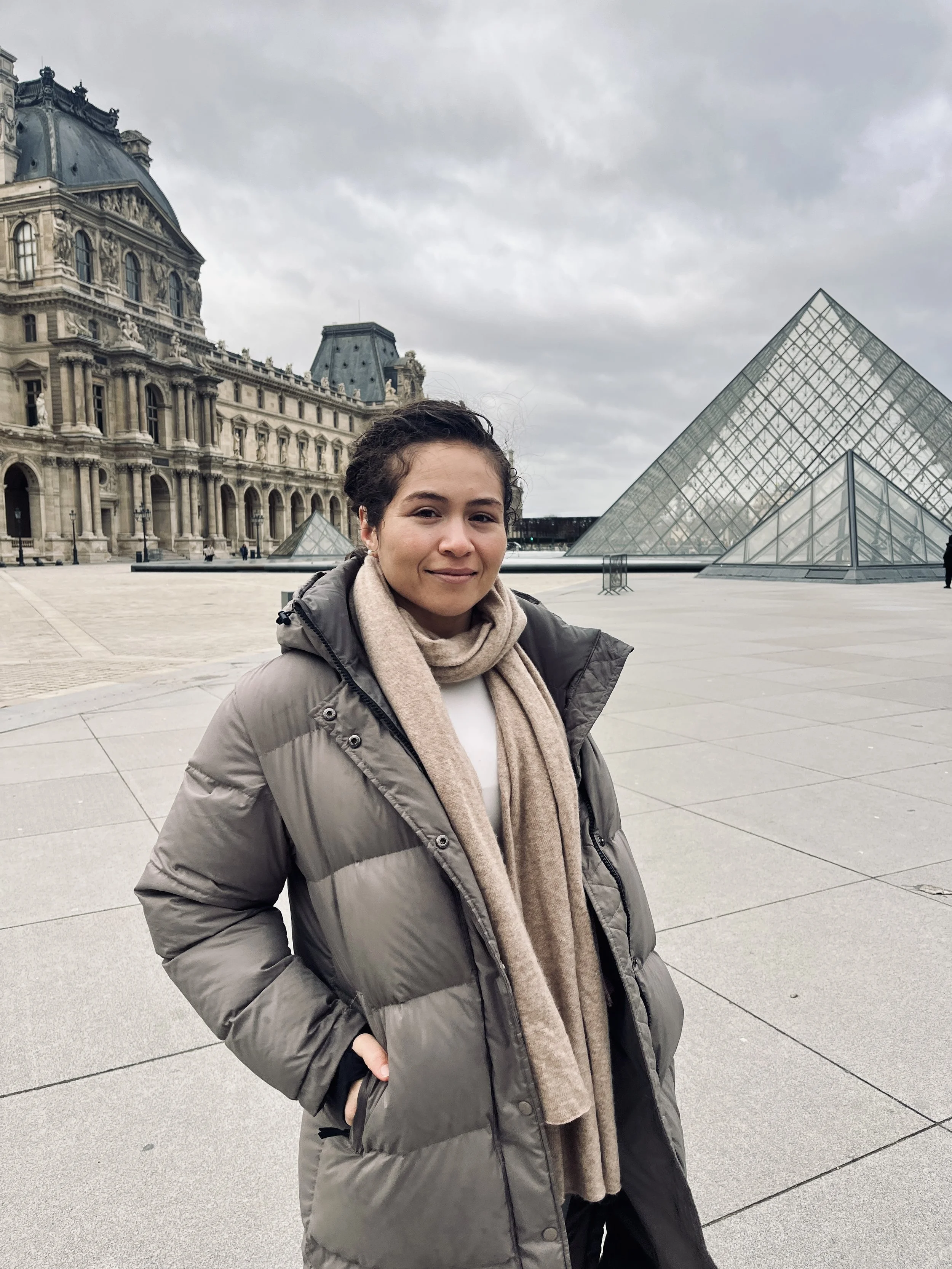 A woman standing in front of the Louvre Museum and glass pyramids in Paris, France, on a cloudy day.